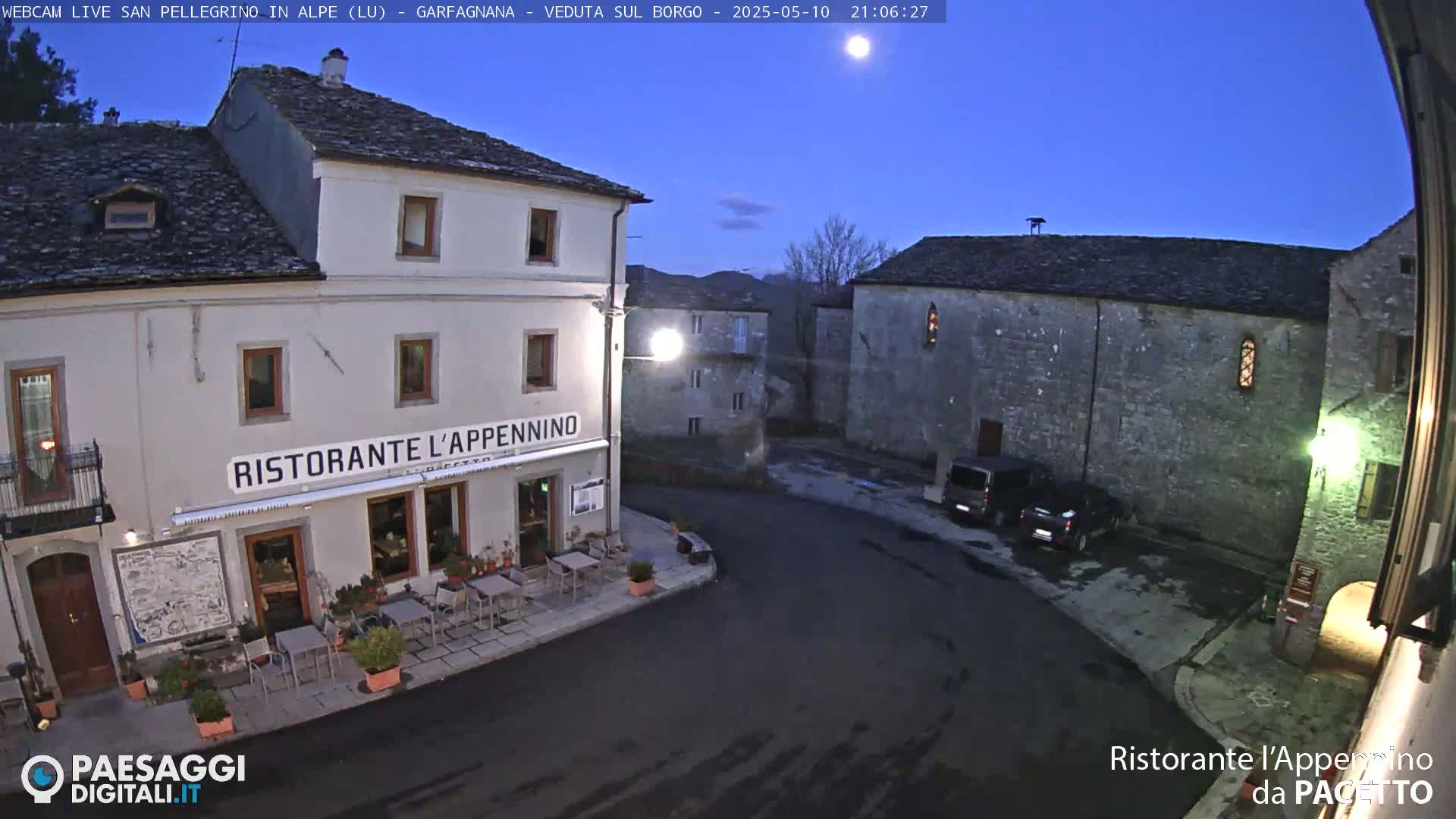 Under a twilight sky, a town square features stone buildings, a restaurant with outdoor seating, and a few parked cars.
