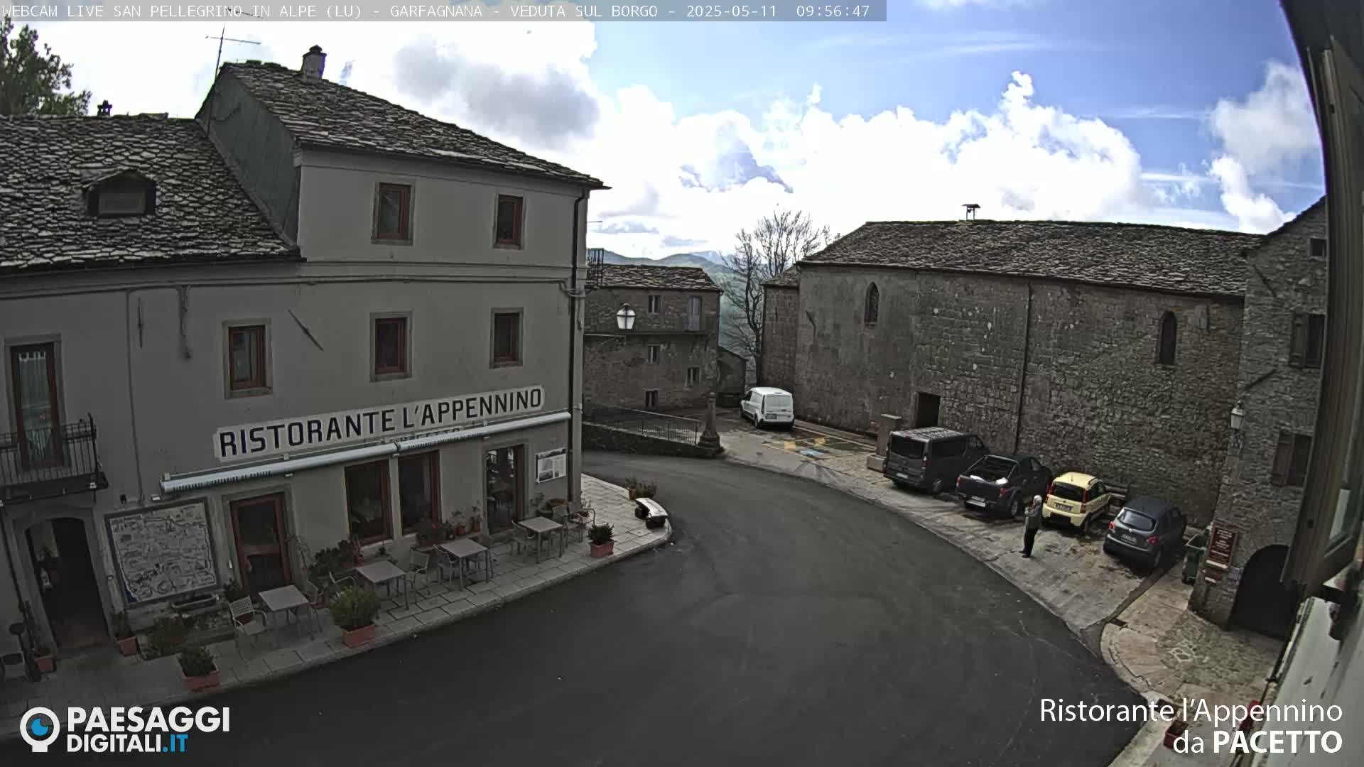 A partly cloudy day reveals a quaint town square with several parked cars, a restaurant, and stone buildings.