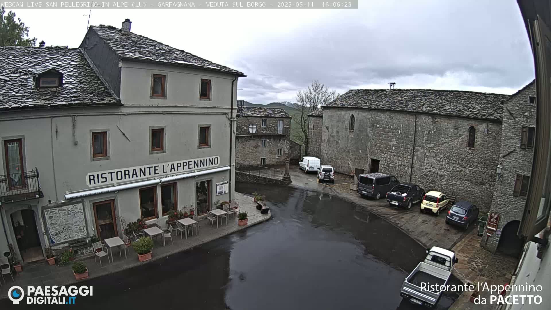 A wet, overcast town square with several parked cars and a restaurant with outdoor seating.