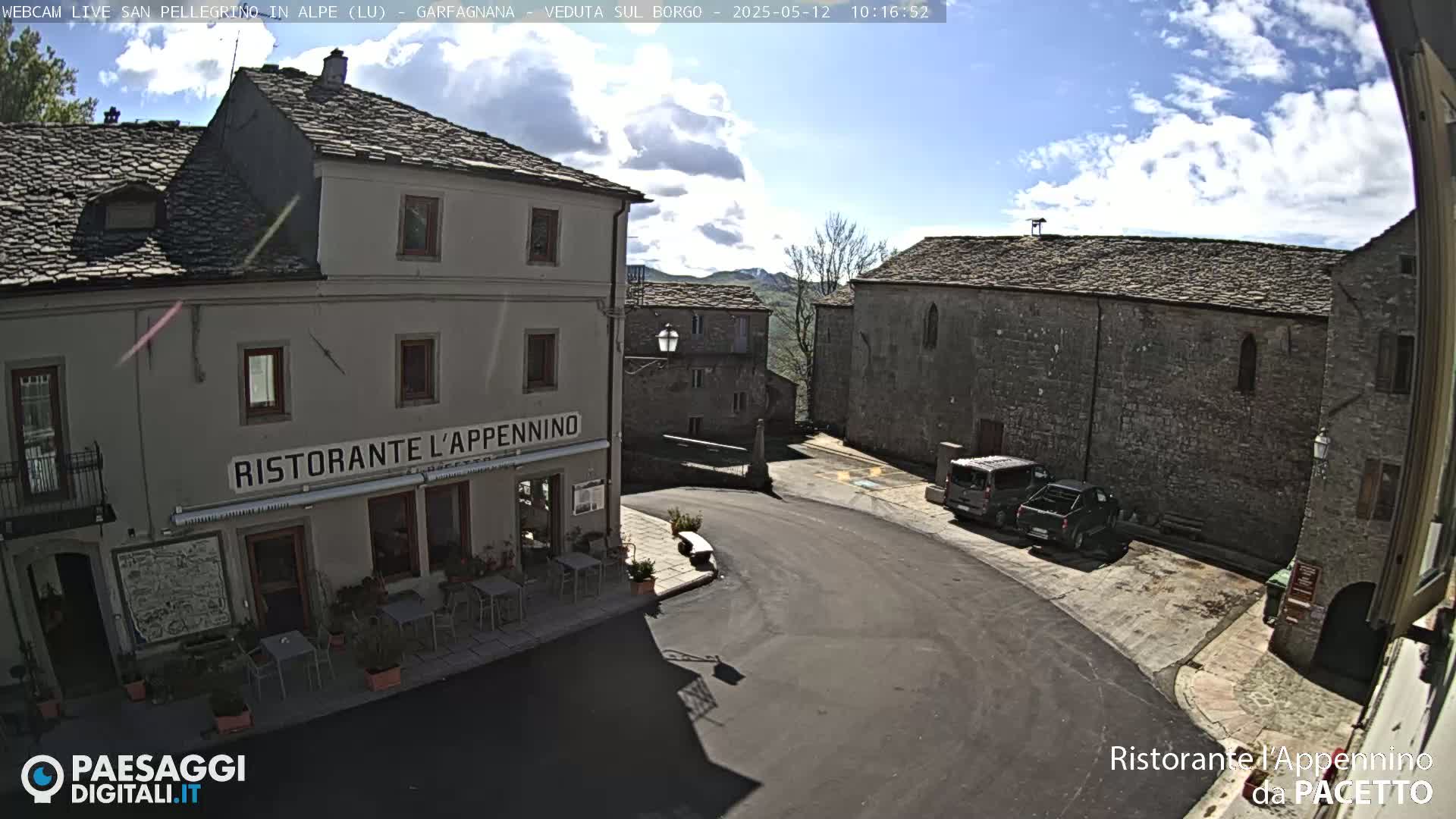 A sunny day shows a town square with parked vehicles in front of stone buildings, one of which is a restaurant.