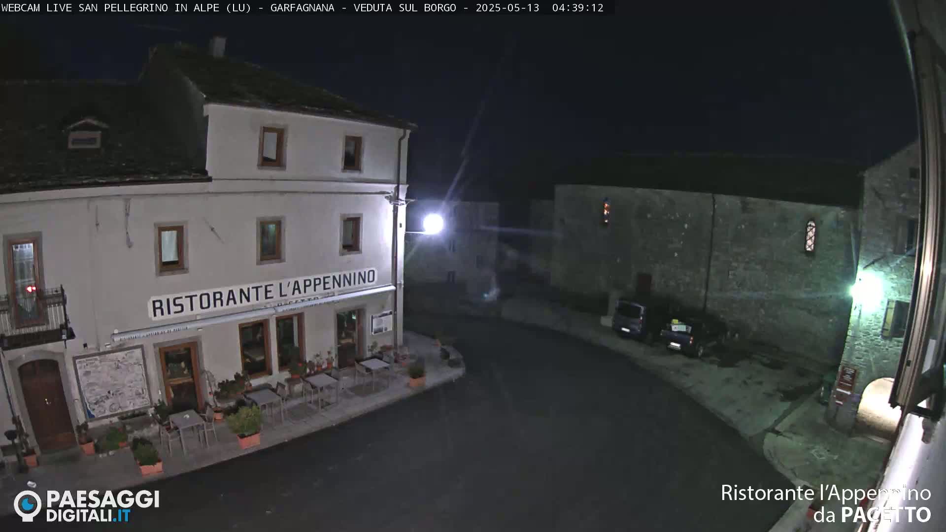 A nighttime, outdoor view of a town square with a restaurant, some parked cars, and old stone buildings under a dark sky.
