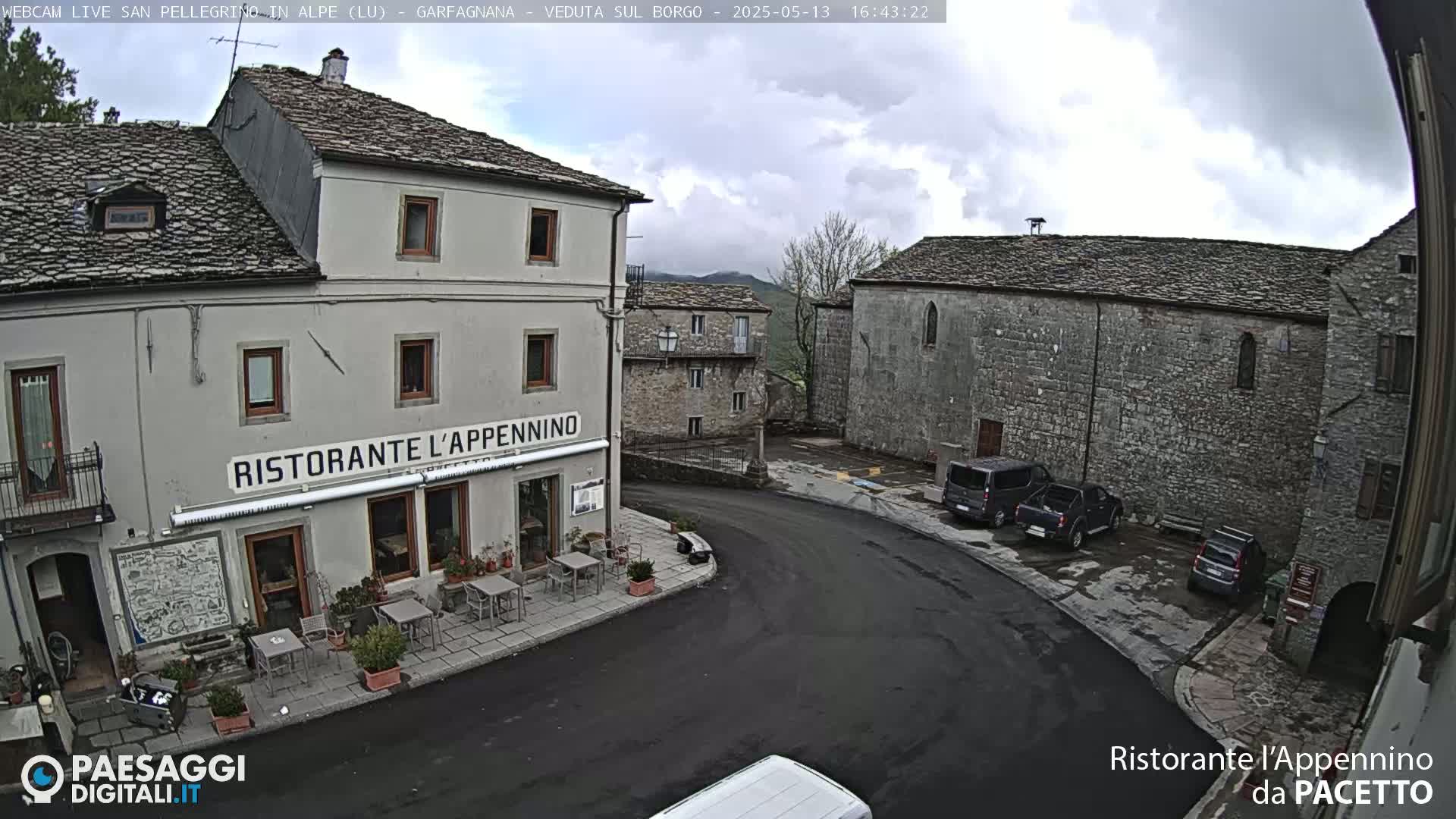 A stone village square on an overcast day with several parked vehicles near a restaurant with outdoor seating.