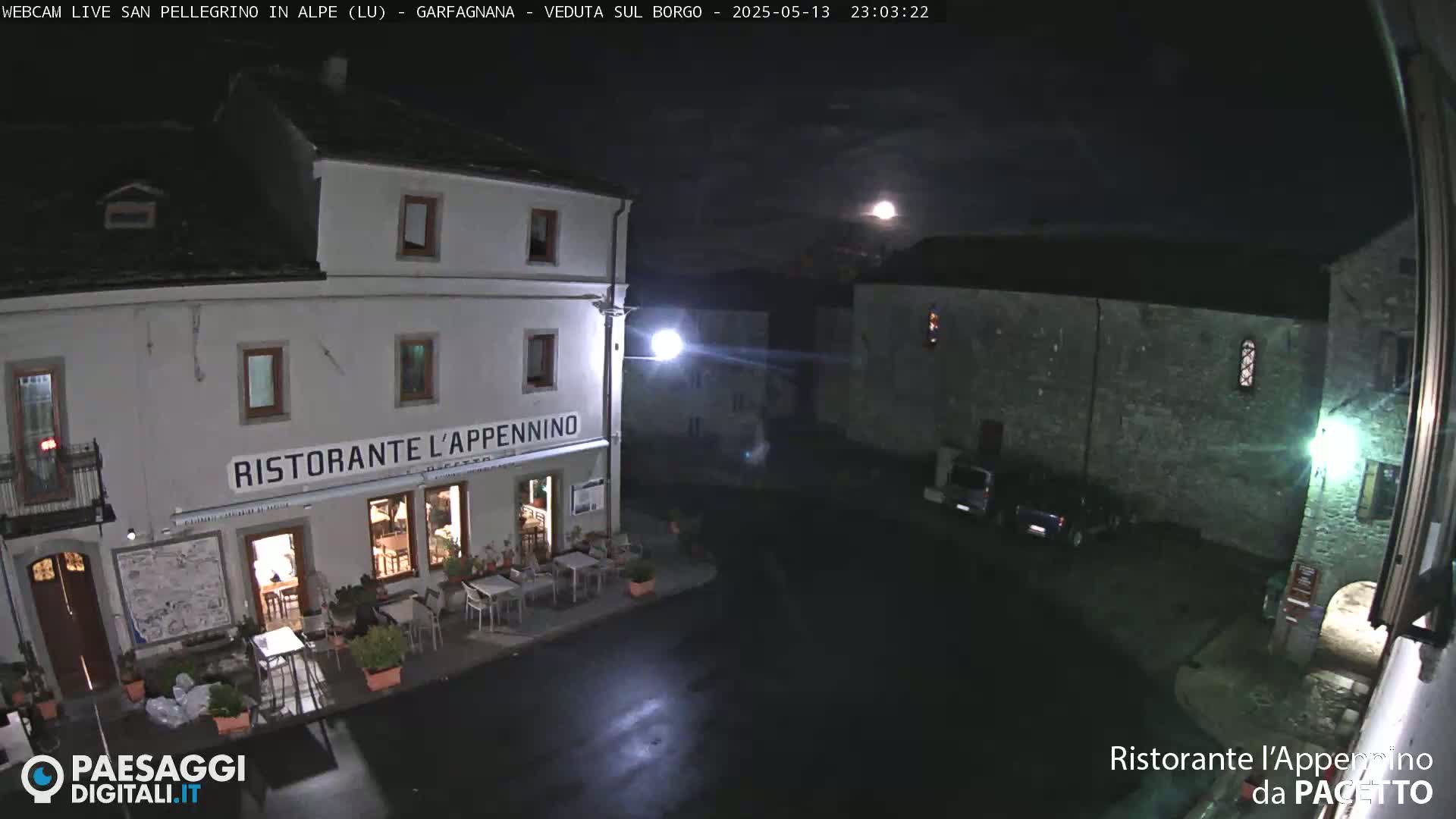 A nighttime view of a town square with a restaurant, illuminated by streetlights and a full moon under a mostly clear sky.