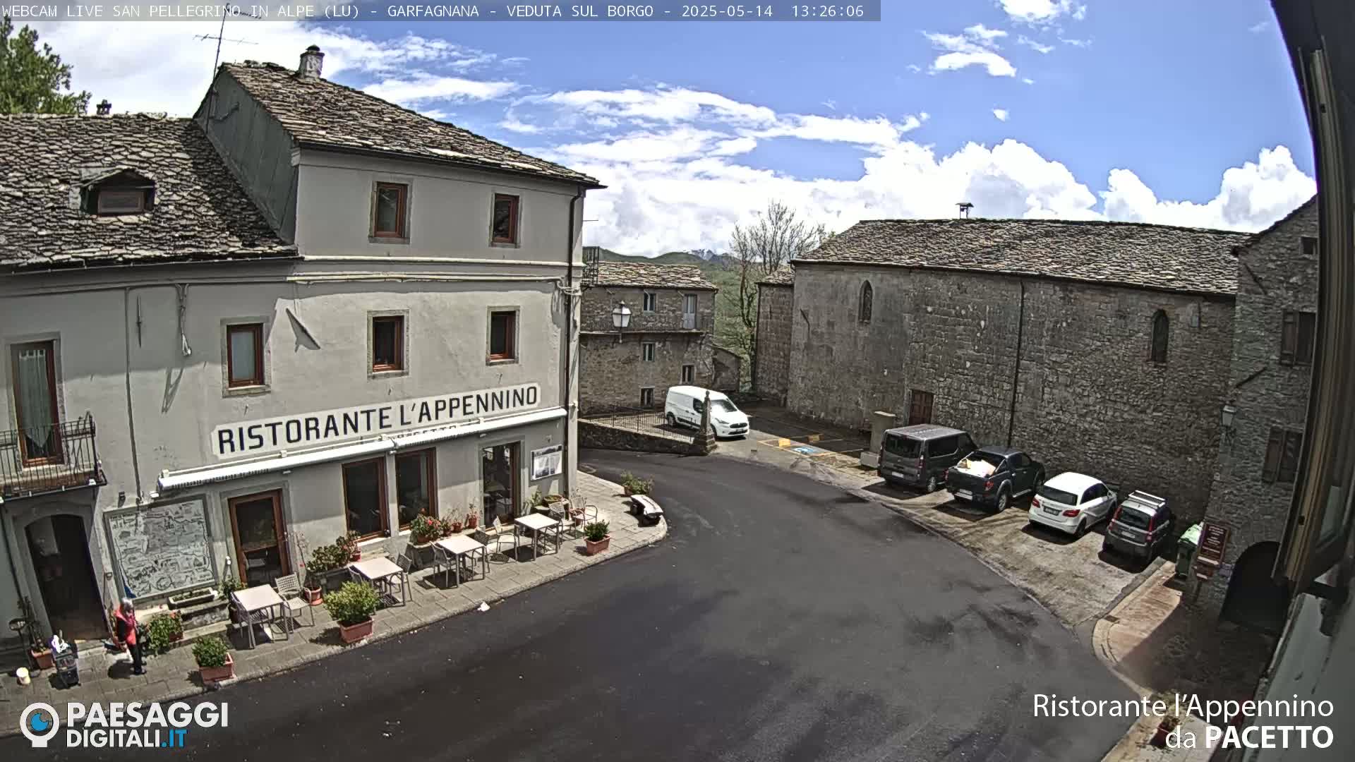 A partly sunny day shows a village square with stone buildings, a restaurant with outdoor seating, and several parked cars.