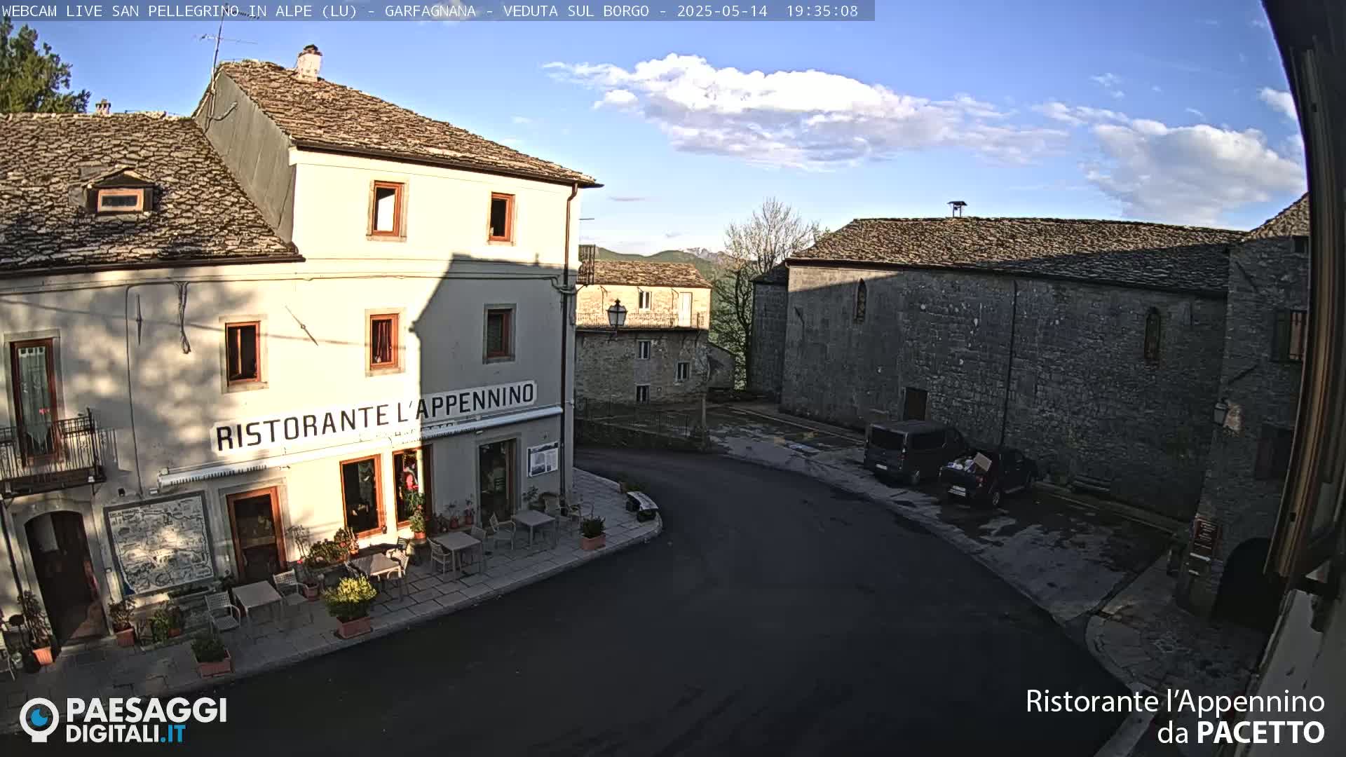 A partly sunny view of a quaint stone village square, with a restaurant and parked cars, under a mostly clear sky.