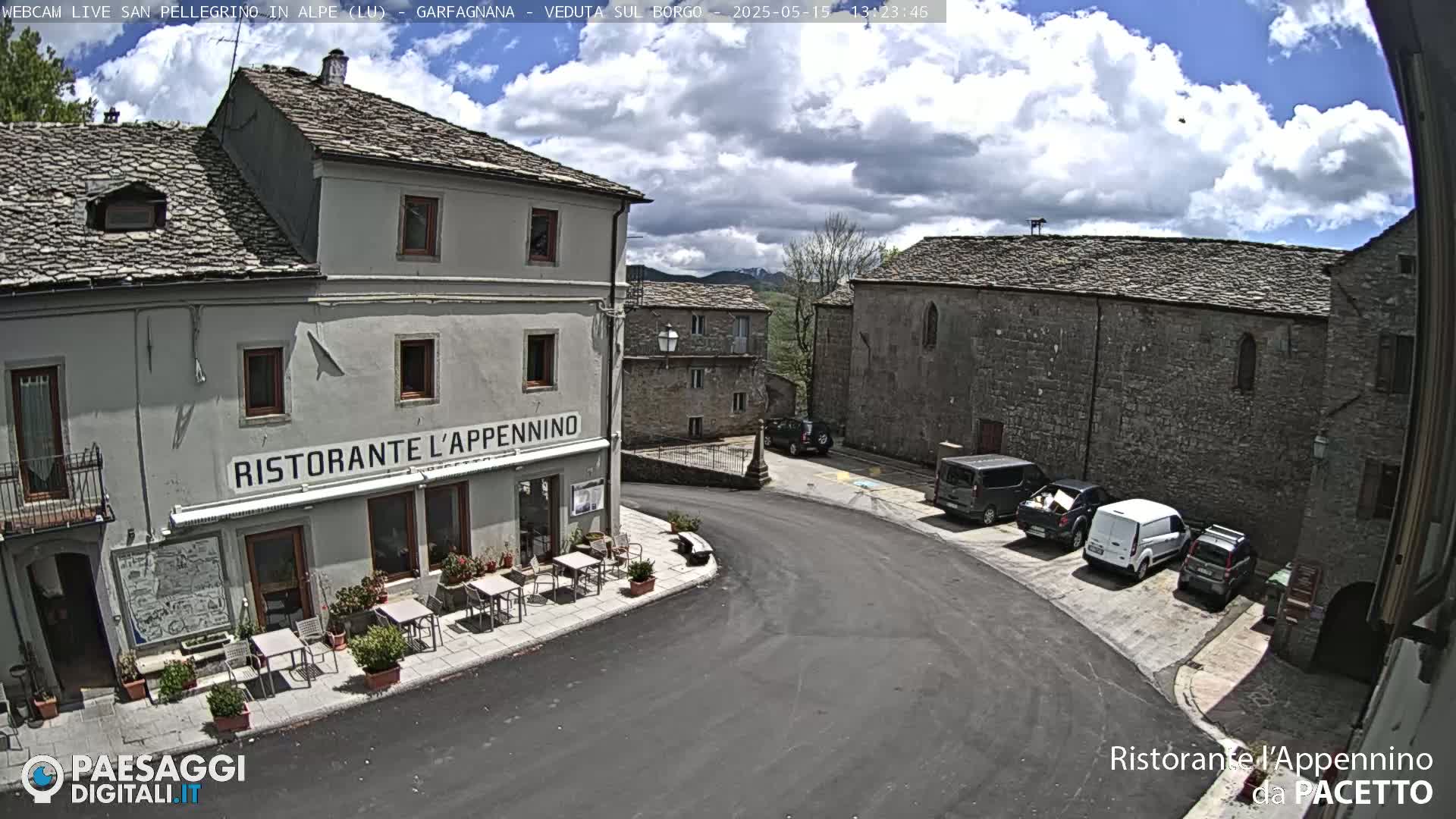 A partly cloudy day reveals a quaint village square with a restaurant, several parked vehicles, and stone buildings under a partly cloudy sky.