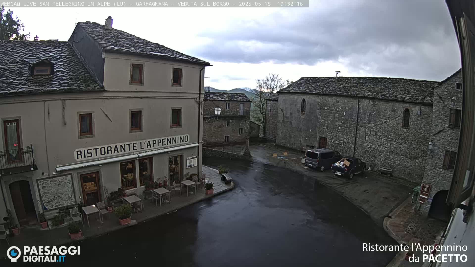 A wet, overcast town square in a small village, featuring a restaurant with outdoor seating and several stone buildings, with a few vehicles parked nearby.