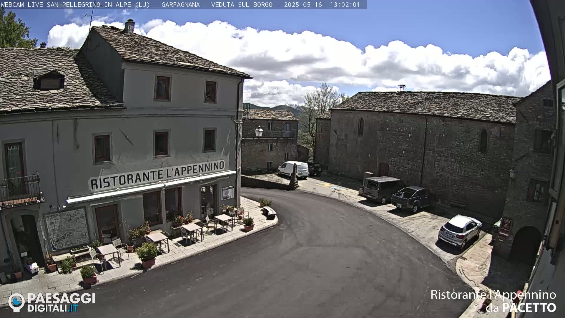 A partly sunny day shows a village square with several parked cars, and a restaurant with outdoor seating.