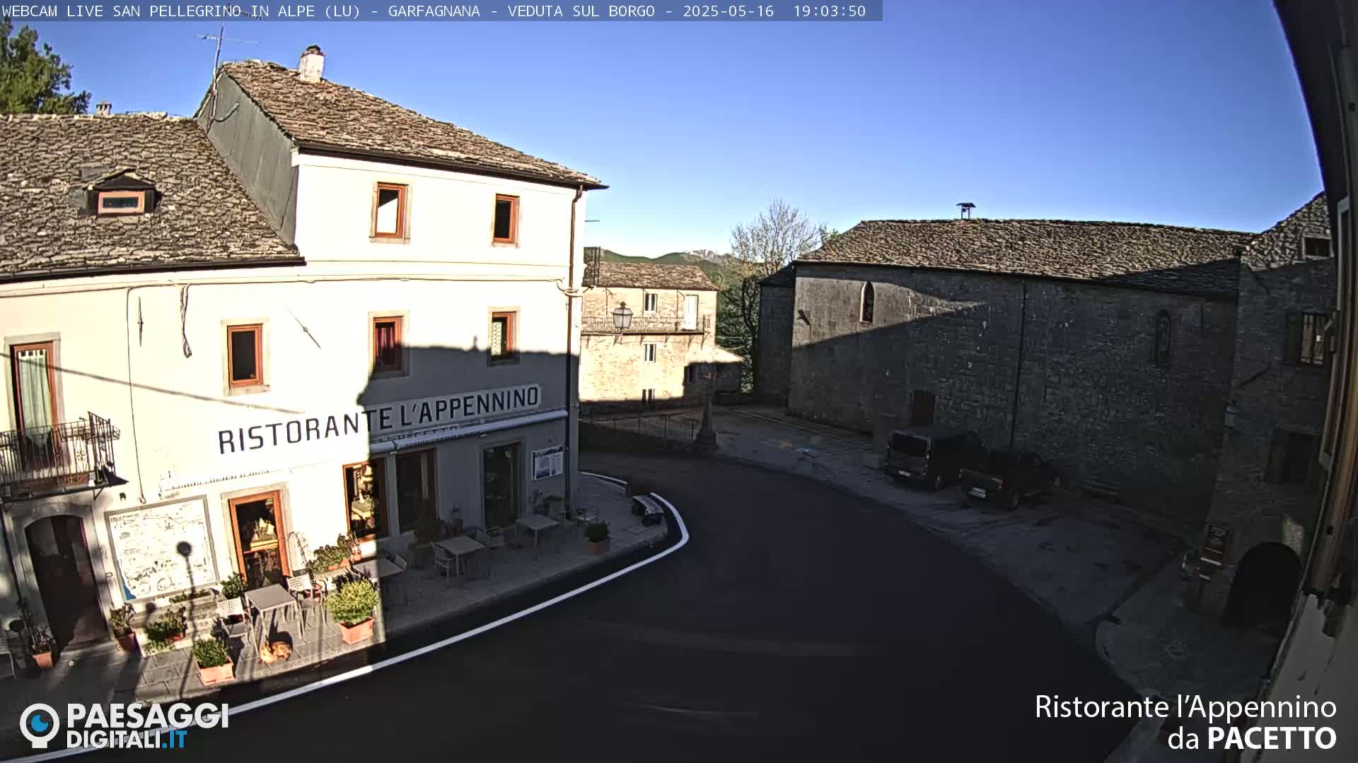 A sunny day reveals a quaint village square with stone buildings, one of which houses a restaurant, and a few parked cars.