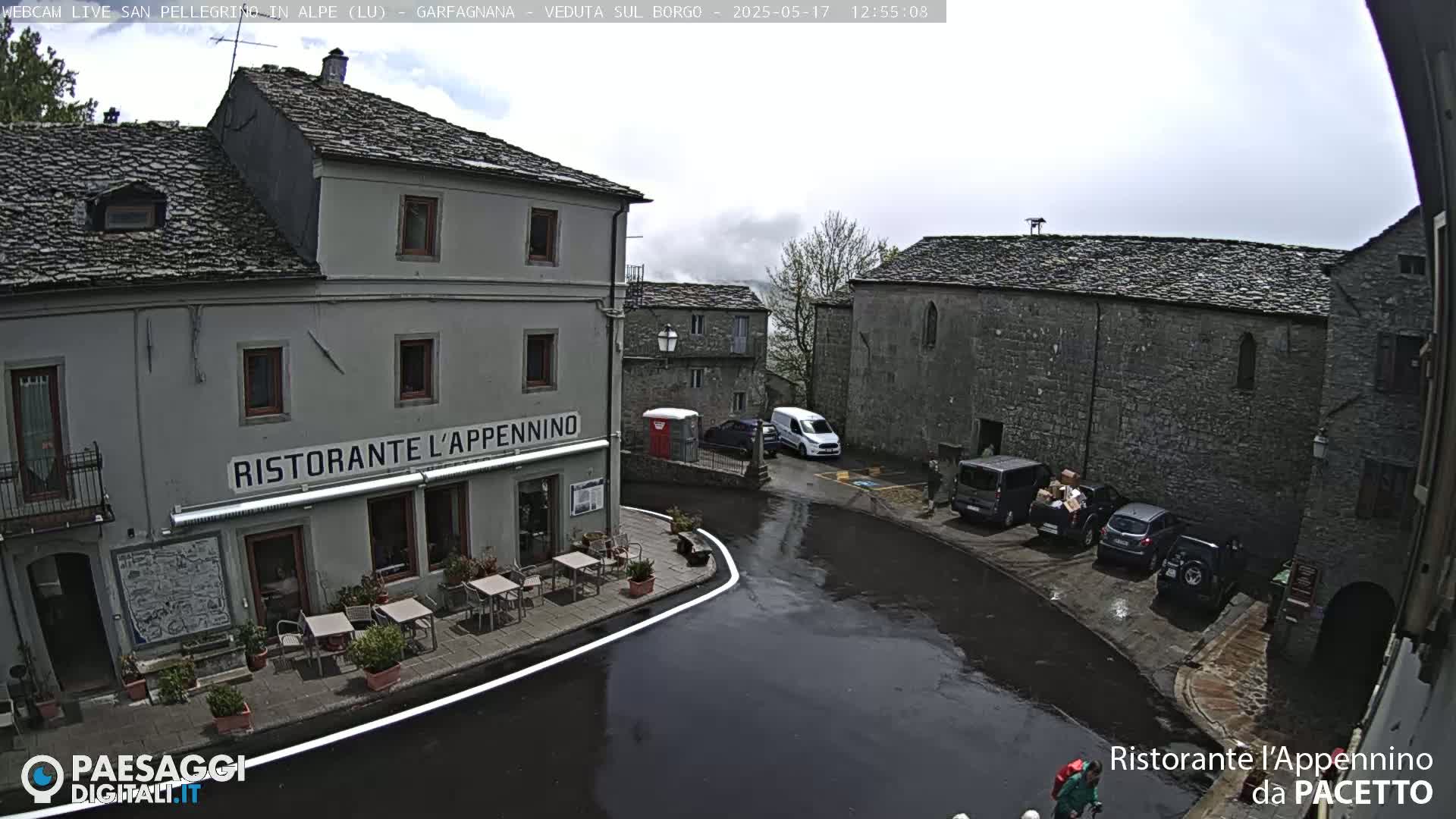 A wet town square in overcast conditions features several parked vehicles near a restaurant with outdoor seating.