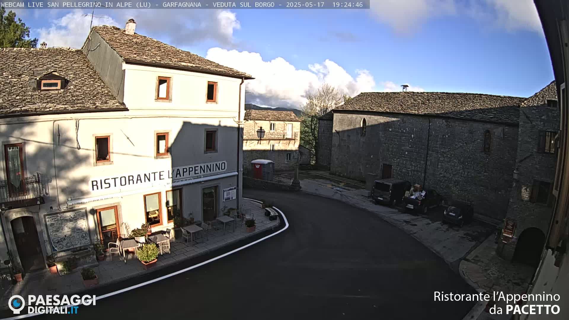 A partly cloudy afternoon scene shows a village square with stone buildings, including a restaurant with outdoor seating, and several parked cars.