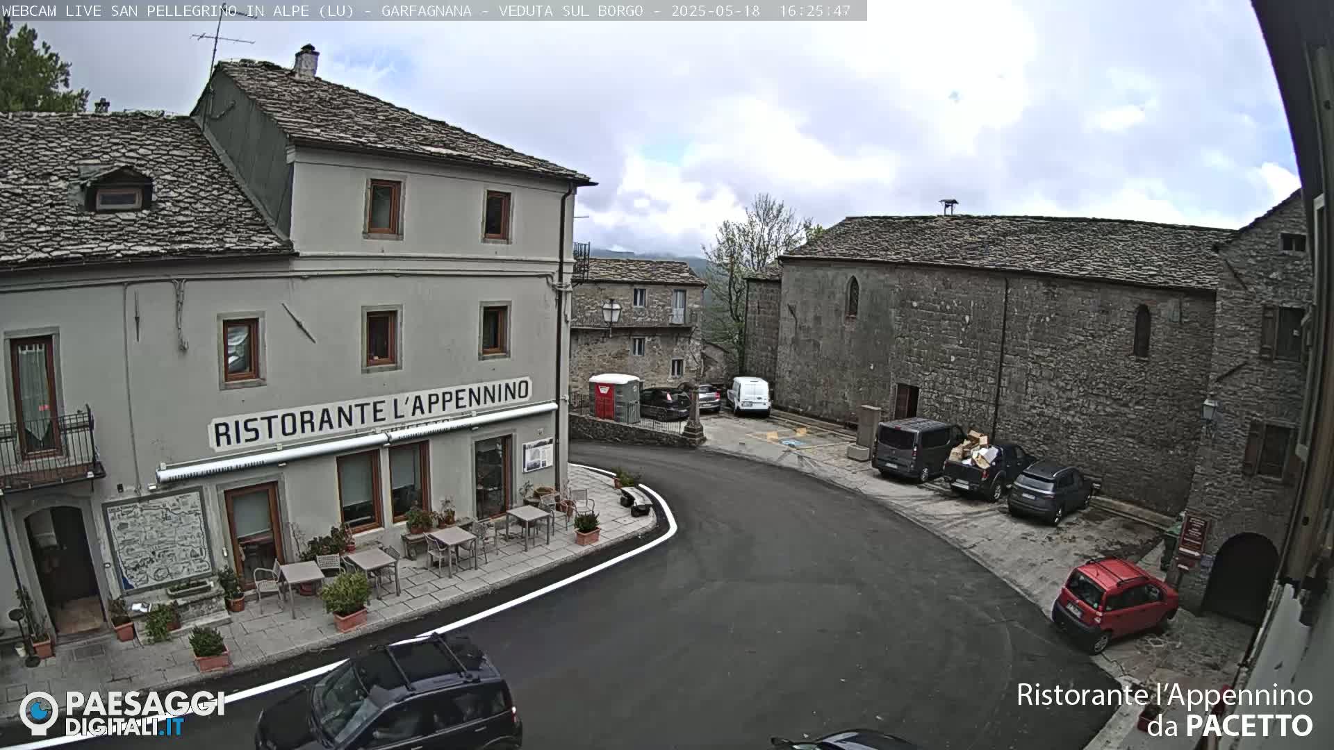 A partly cloudy day reveals a village square with several cars parked near stone buildings, one of which is a restaurant.