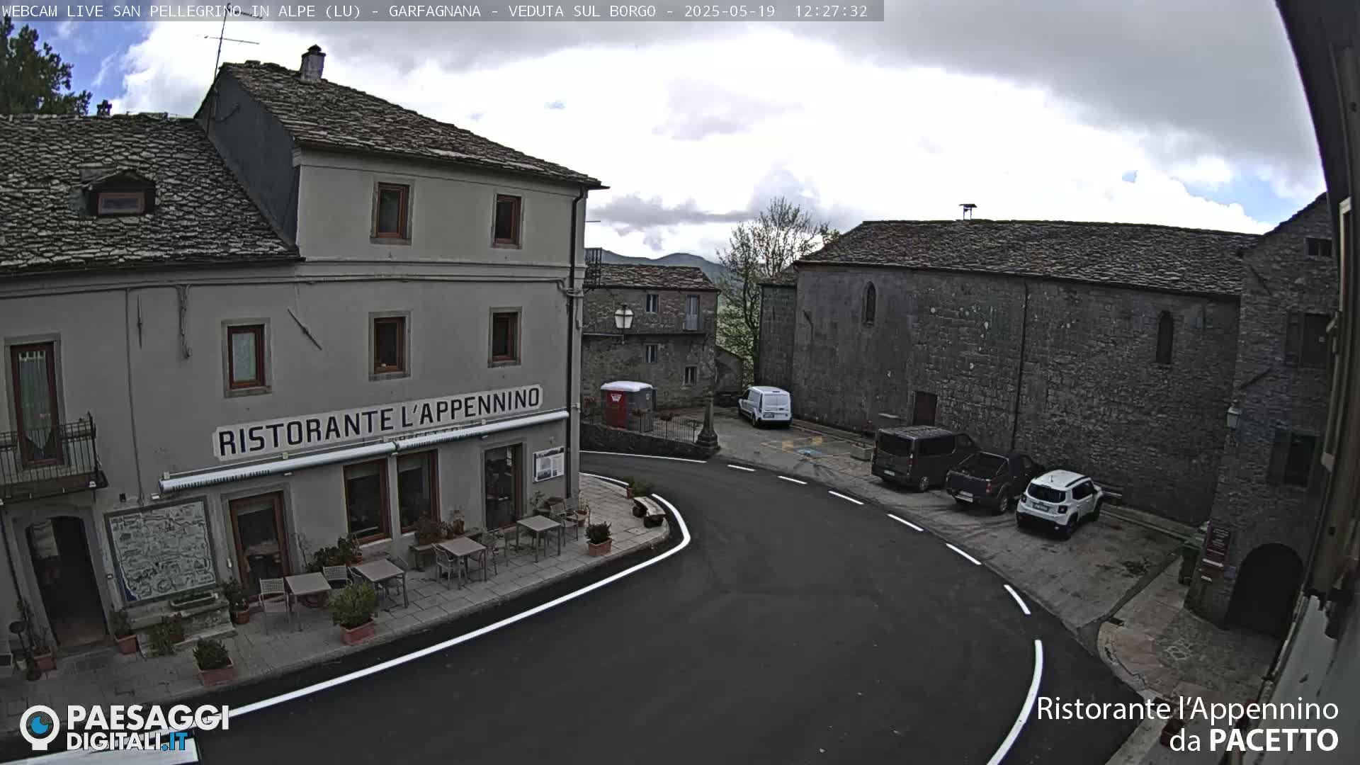 A mostly cloudy day reveals a small town square with several parked cars and a restaurant with outdoor seating.