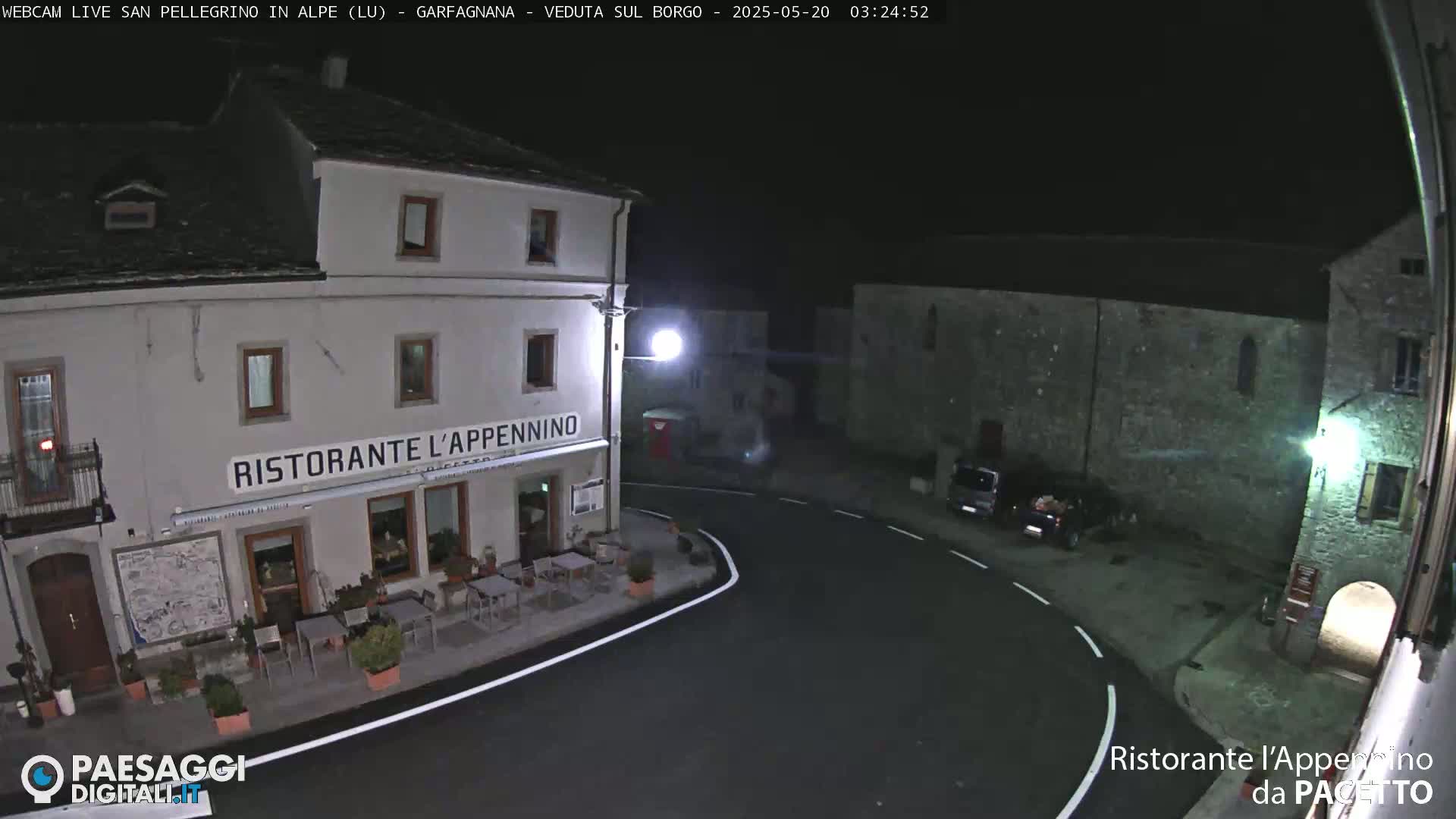 A nighttime view of a quiet town square with a restaurant, several parked cars, and stone buildings under a clear, dark sky.