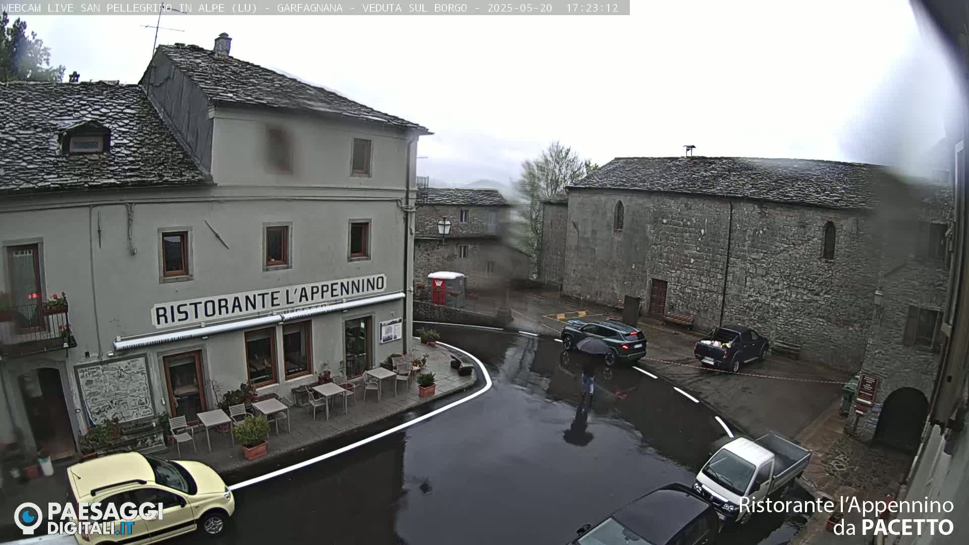 A wet town square with stone buildings, a restaurant with outdoor seating, and several cars parked and driving under a gray, overcast sky.