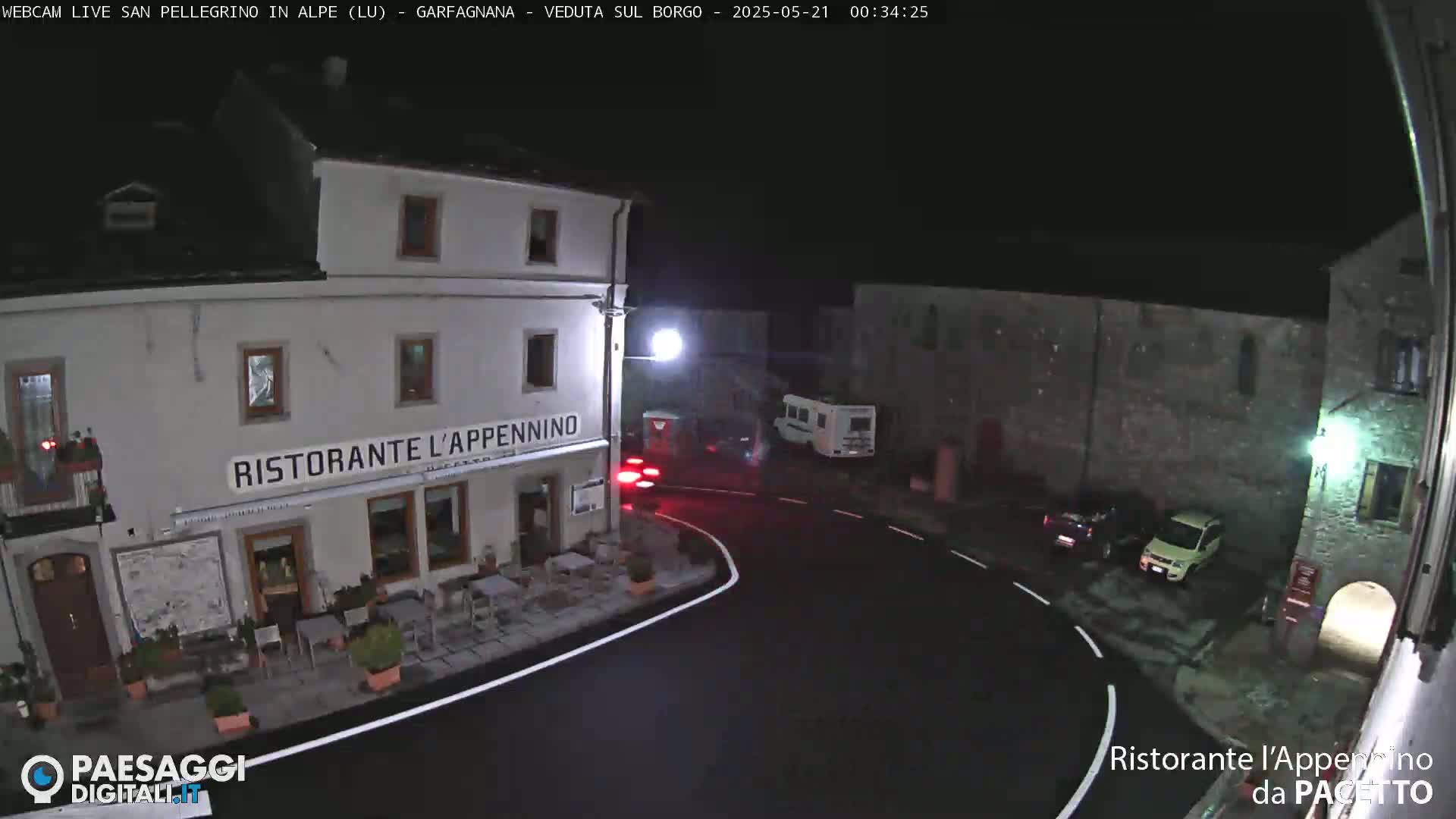 A nighttime view of a village square with a restaurant, parked cars, and a camper van, under clear, dark skies.