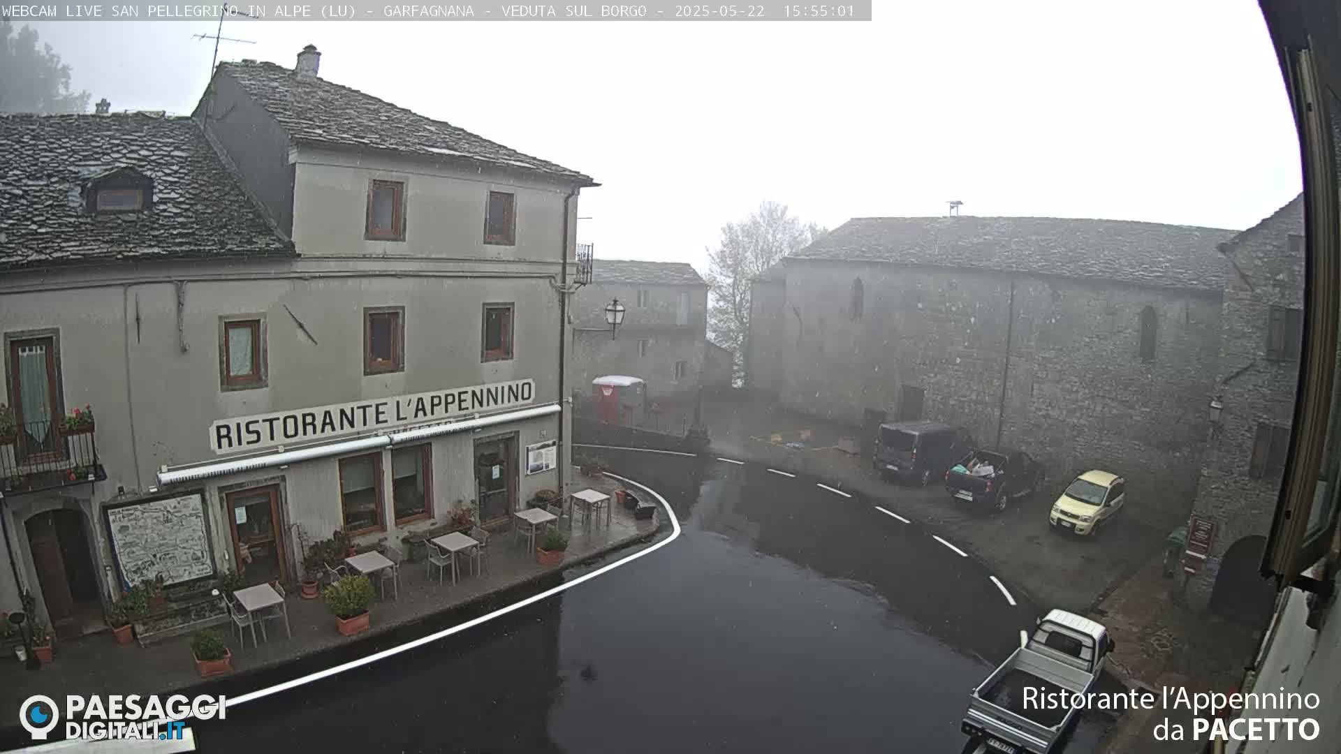 A wet, foggy town square with stone buildings, a restaurant, and several parked cars.