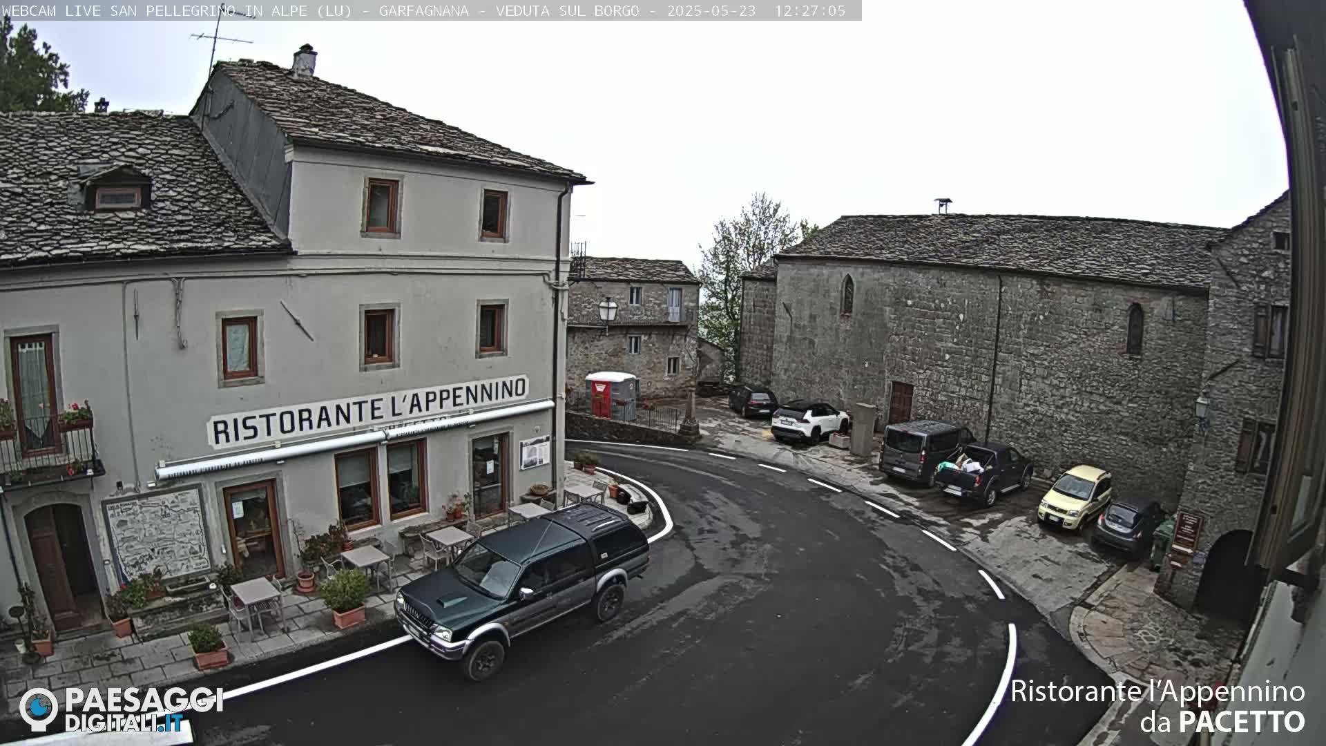 A dark-colored pickup truck is parked in front of a restaurant in a stone village square on an overcast day.