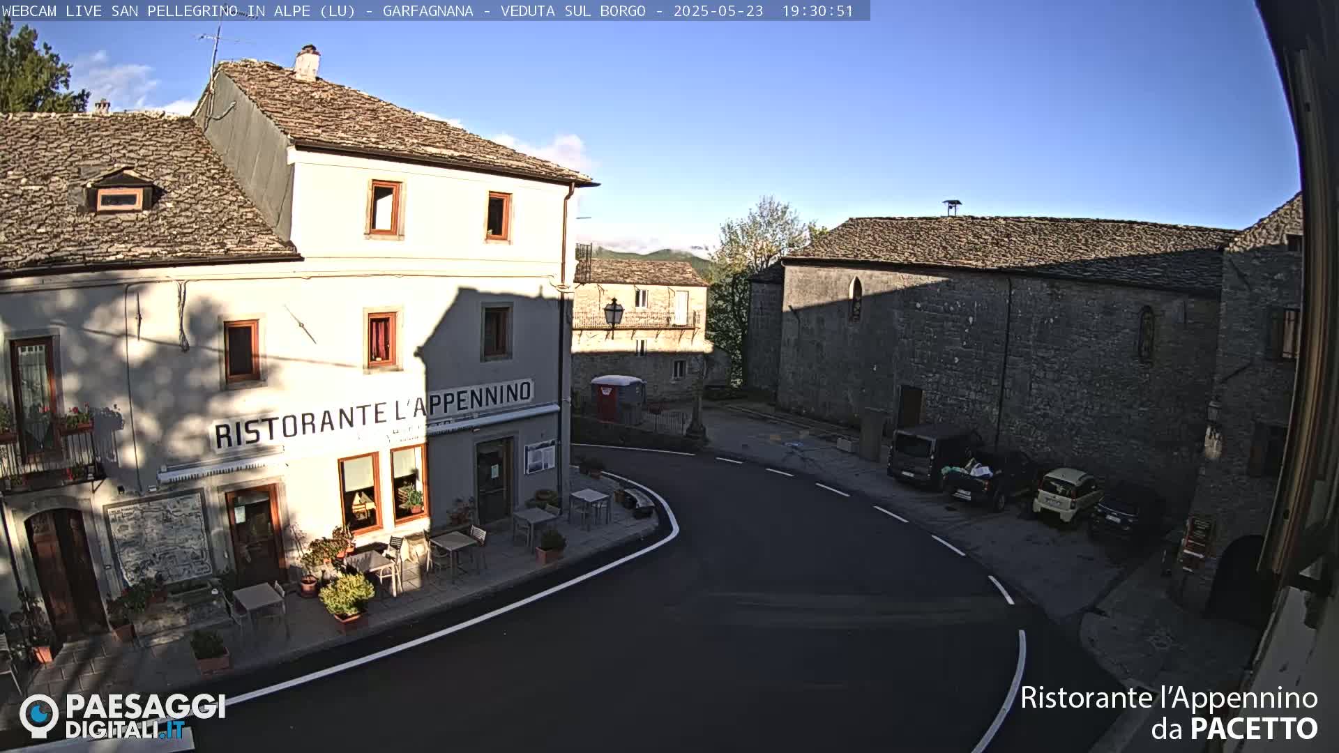 A sunny, clear day reveals a quaint village square with stone buildings, a restaurant, and a few parked cars.