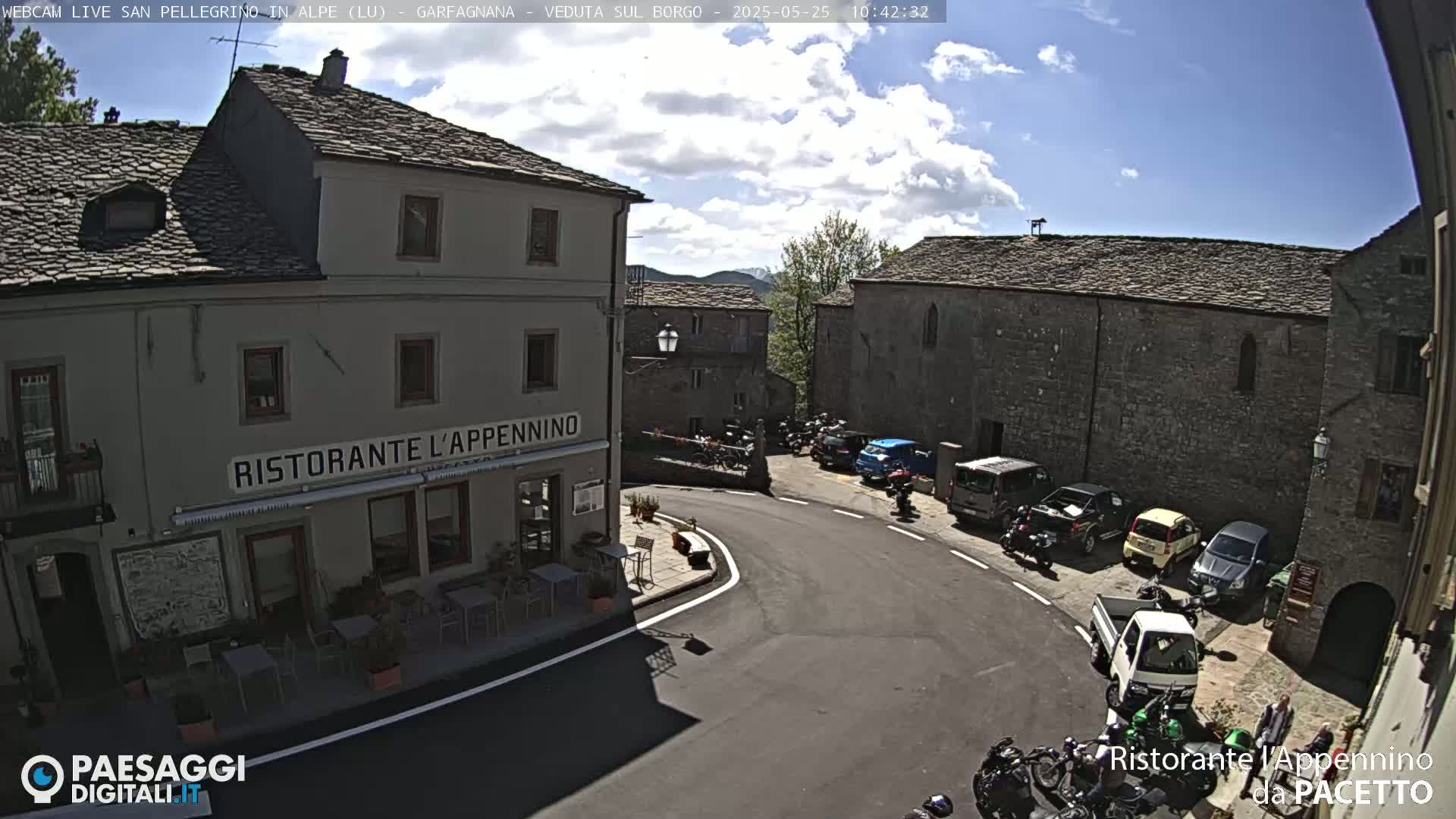 A partly sunny day shows a village square with parked cars and motorcycles surrounding a restaurant.