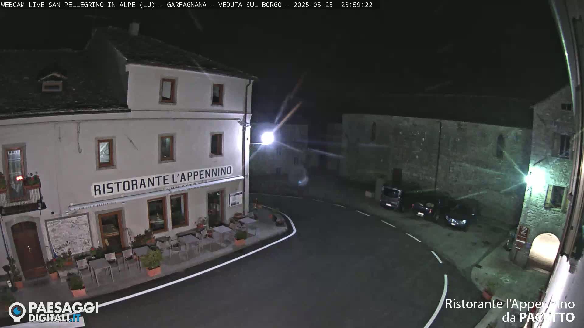 A nighttime, high-angle view of a village square with a restaurant, several parked cars, and stone buildings under a clear, dark sky.