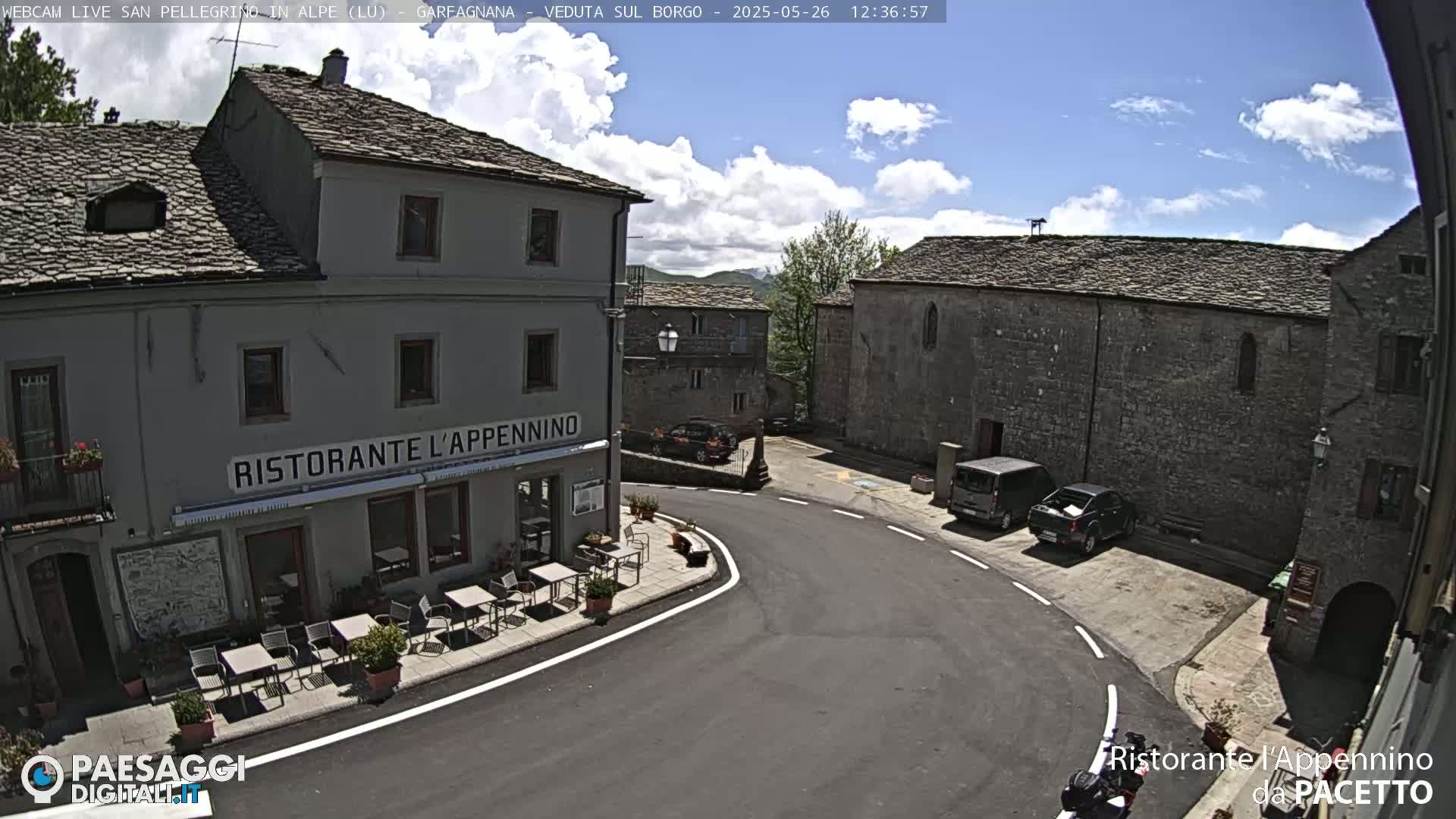 A partly cloudy sunny day reveals a quaint village square with a restaurant, stone buildings, and a few parked vehicles.