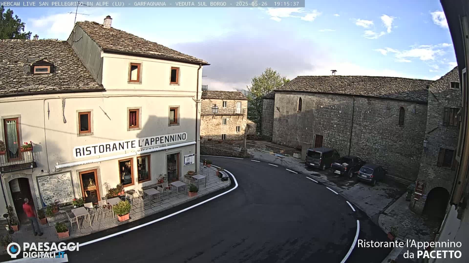 A partly cloudy day in a small village square with stone buildings, a restaurant with outdoor seating, and several parked vehicles.