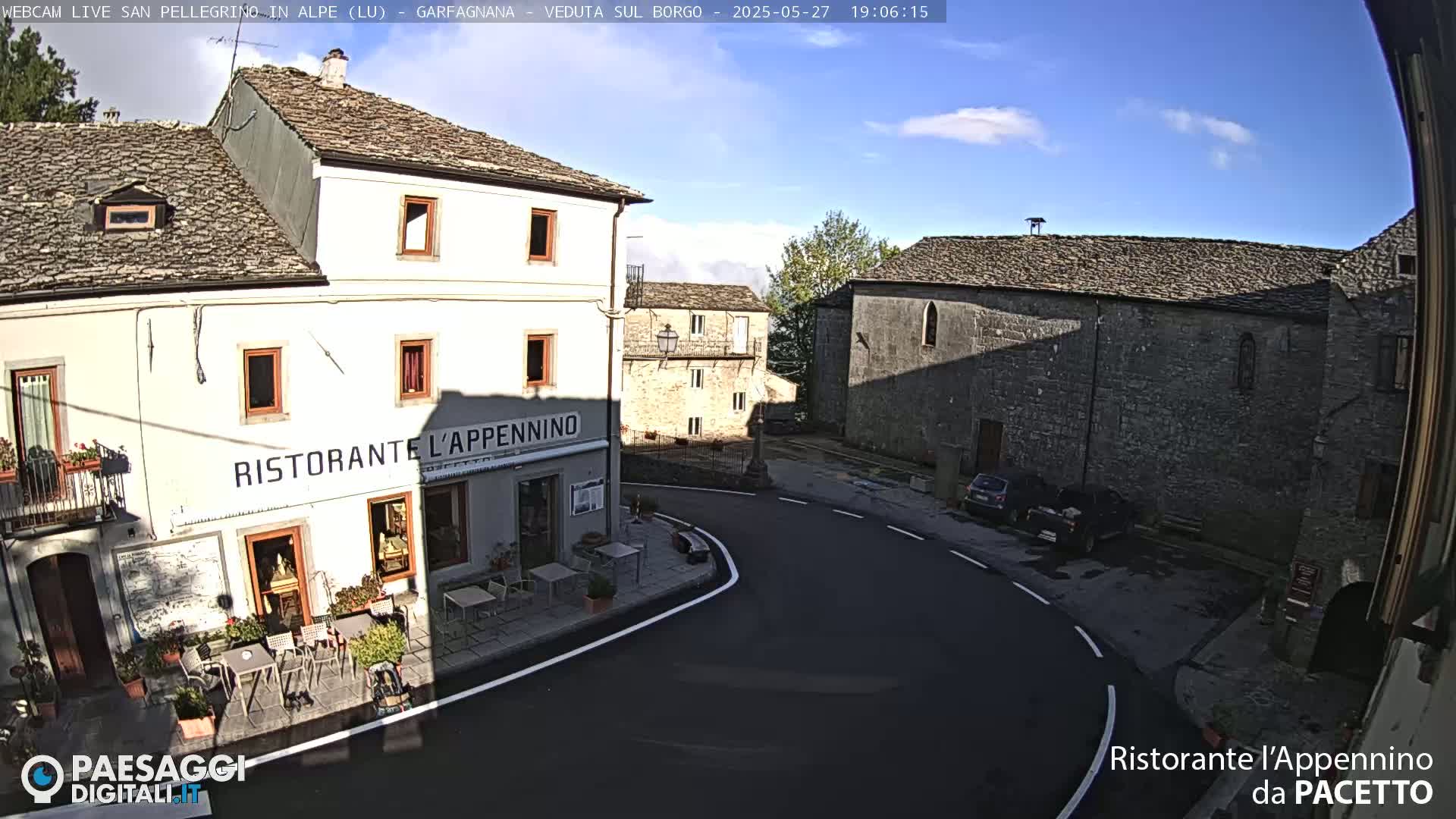 A sunny, outdoor view of a quaint village square with stone buildings, one of which houses a restaurant, and a few parked cars.