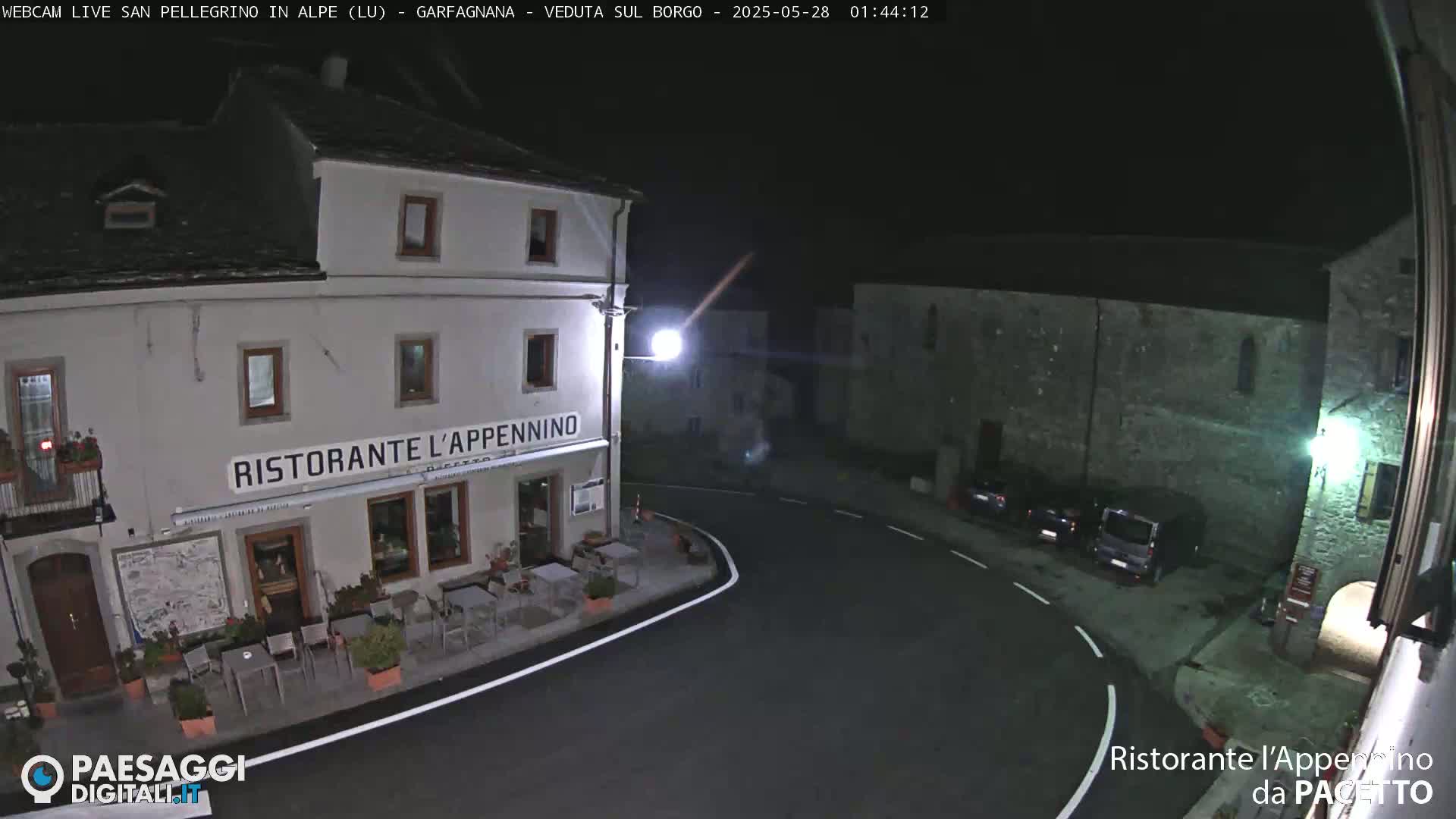 A nighttime view of a town square with a restaurant, several parked cars, and stone buildings under a clear, dark sky.