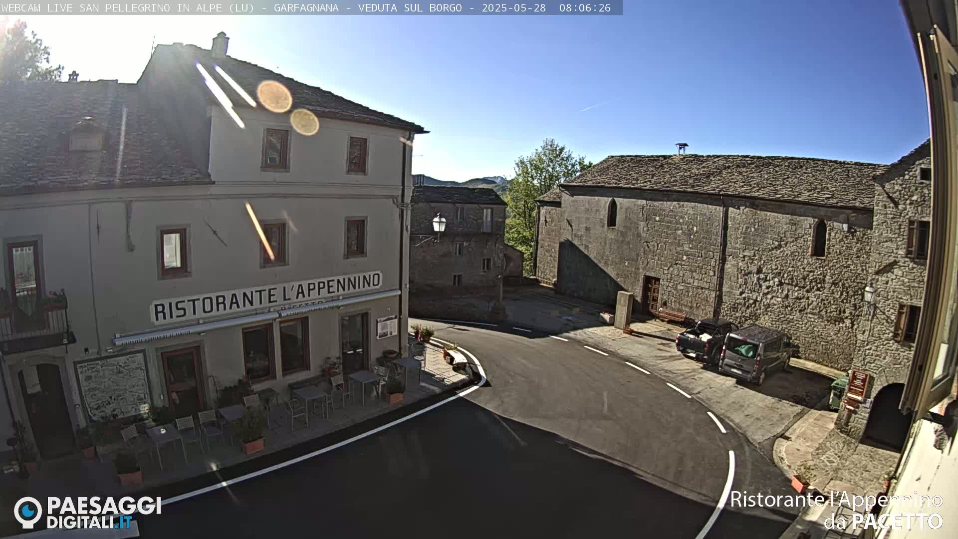 A sunny view of a town square with stone buildings, one of which is a restaurant, and a few parked vehicles.