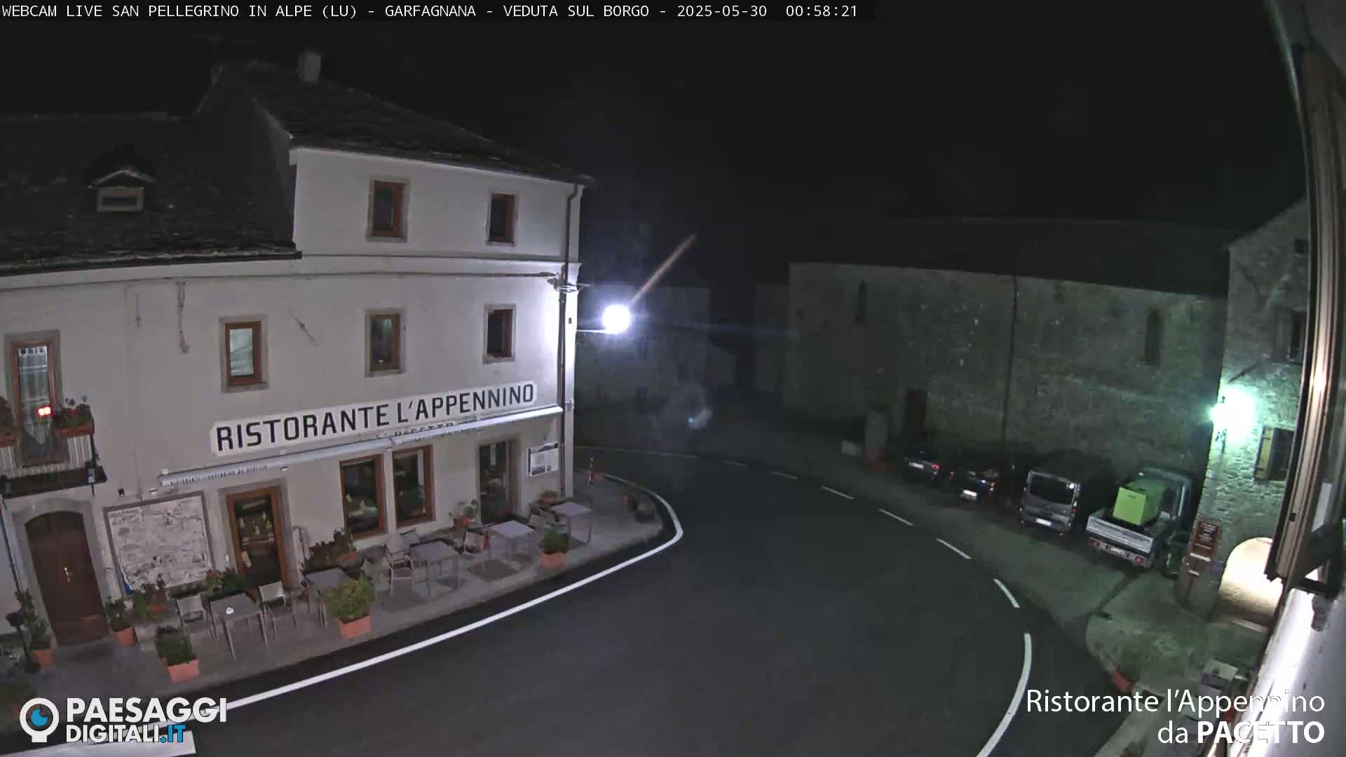 A nighttime, high-angle view of a town square with a restaurant and parked vehicles.