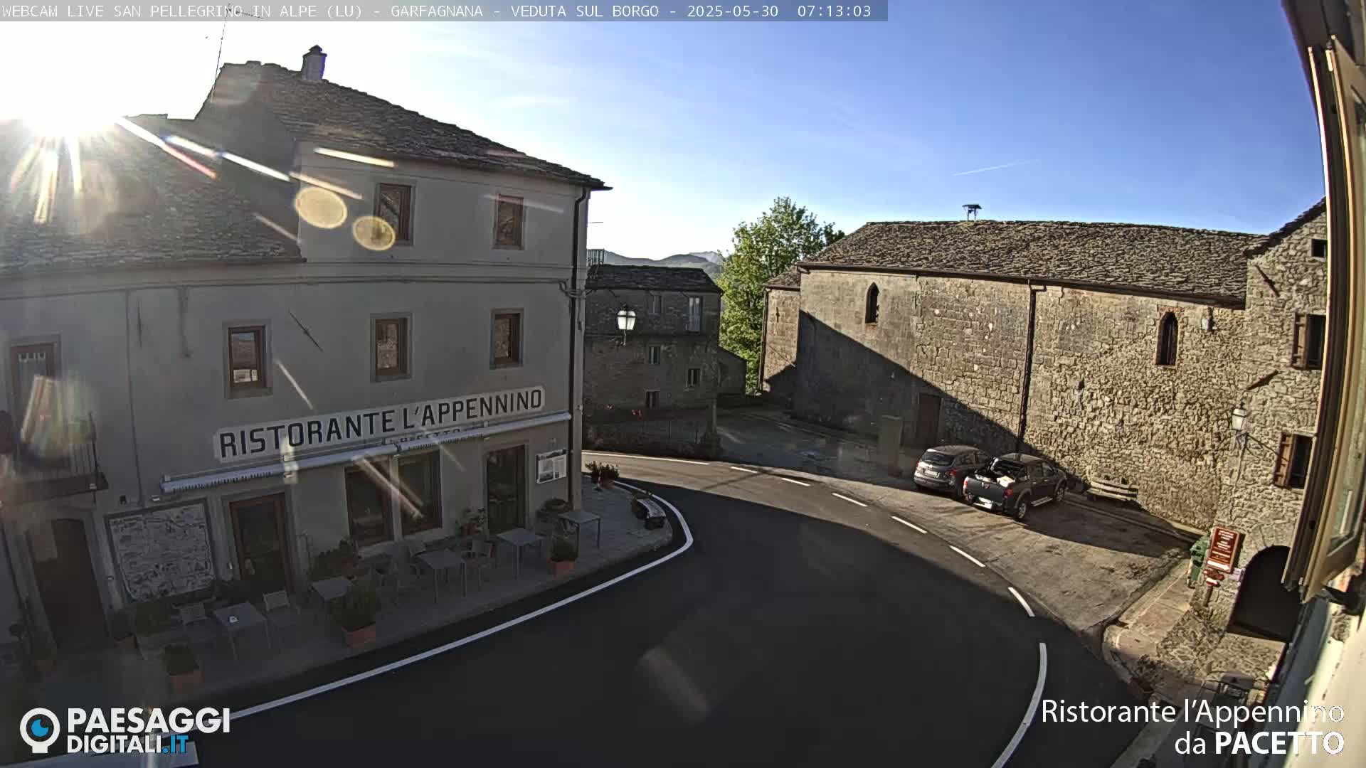 A sunny view of a town square with stone buildings, a restaurant, and a few parked cars.