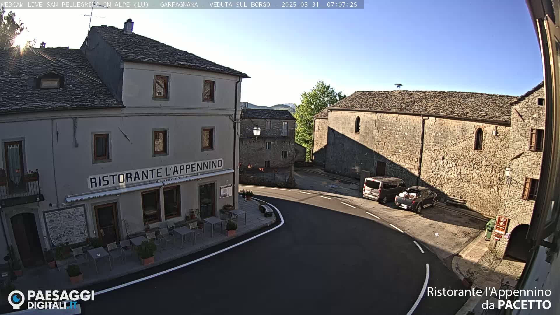 A sunny day reveals a quaint European village square with stone buildings, including a restaurant with outdoor seating, and a few parked vehicles.