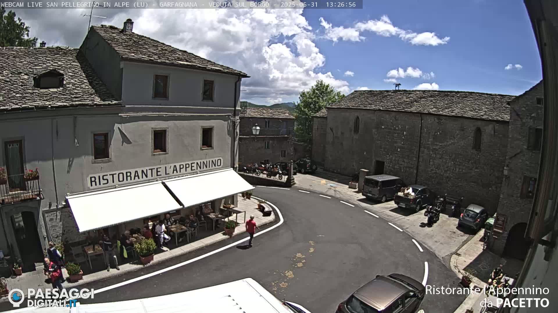A sunny day in a quaint town square with stone buildings, a restaurant with outdoor seating, and parked cars and motorcycles.