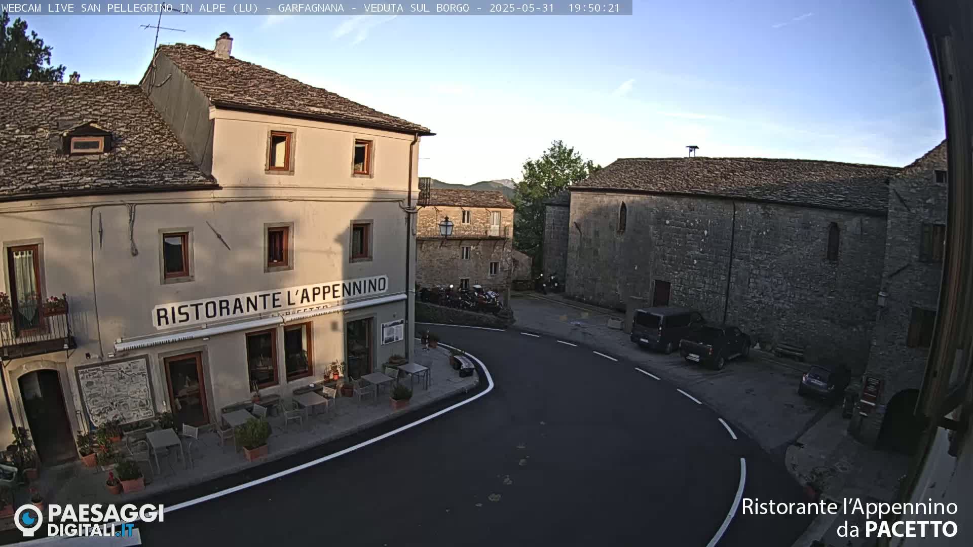 A quaint village square, under a clear evening sky, features stone buildings, a restaurant with outdoor seating, and a few parked vehicles.