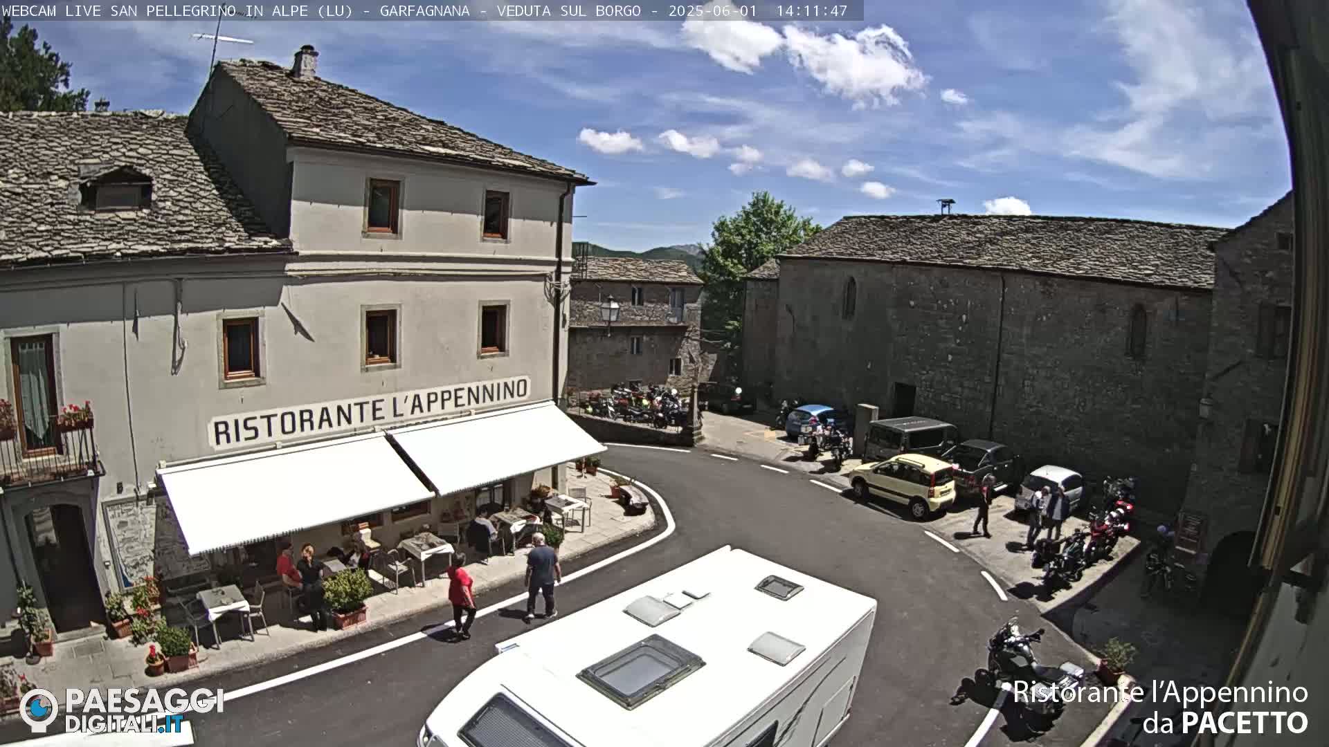 A sunny day reveals a village square with stone buildings, a restaurant with outdoor seating, and several parked cars and motorcycles.