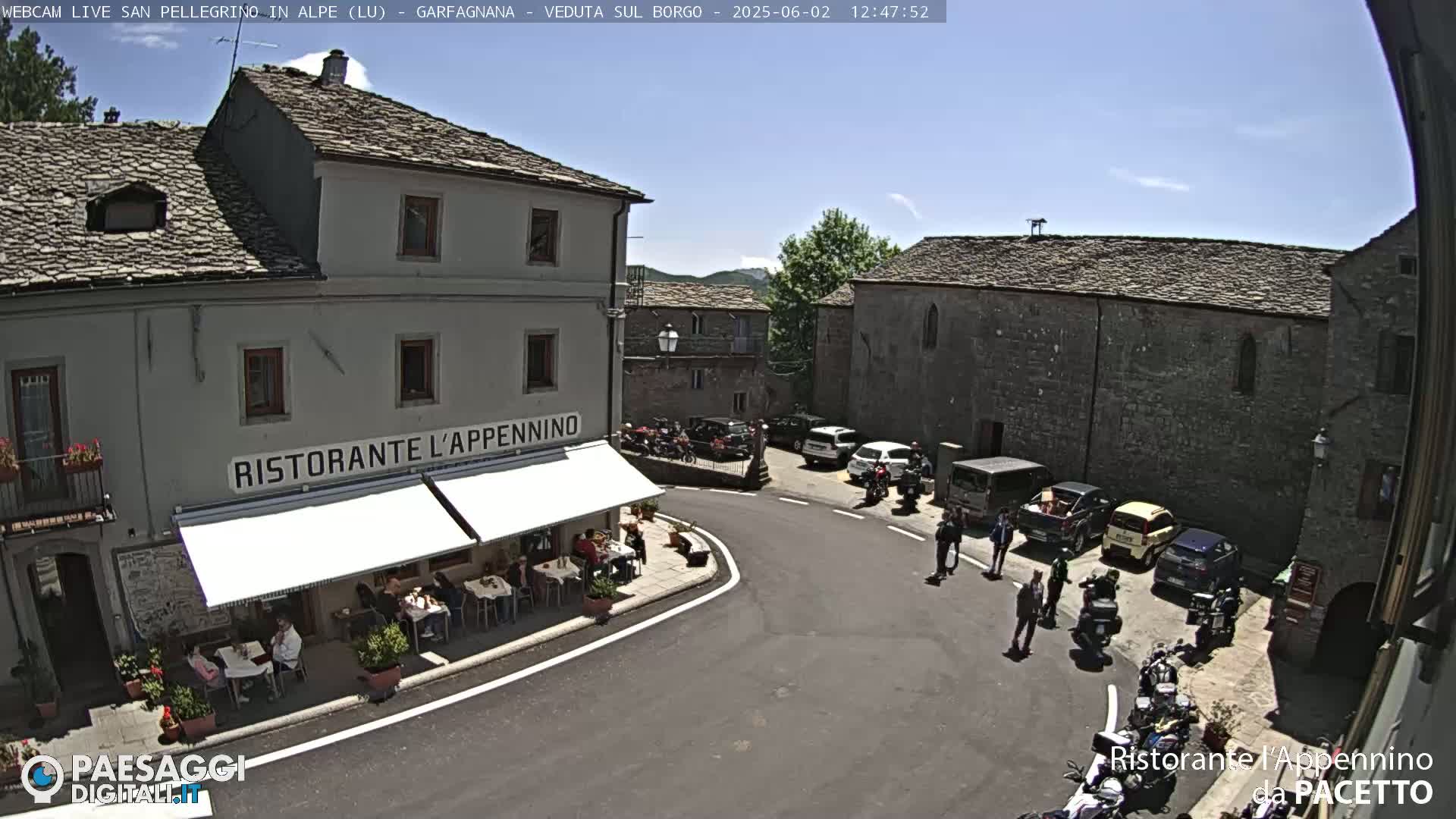 A sunny day in a stone village square shows several parked cars and motorcycles near a restaurant with outdoor seating.