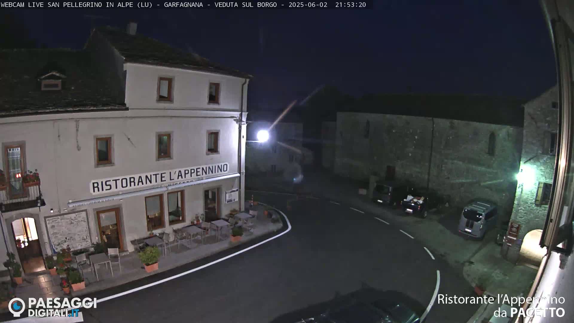 A nighttime view of a village square with a restaurant, several parked cars, and stone buildings under a dark, clear sky.