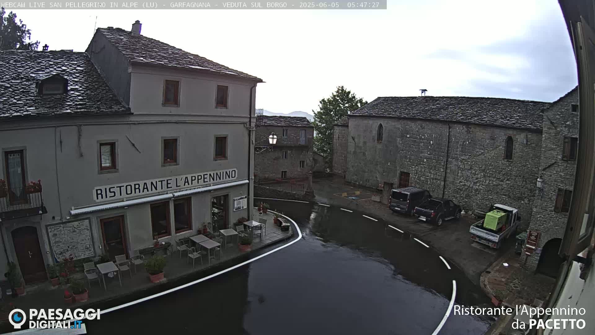A wet village square with stone buildings, one of which is a restaurant, is viewed from above on an overcast day.
