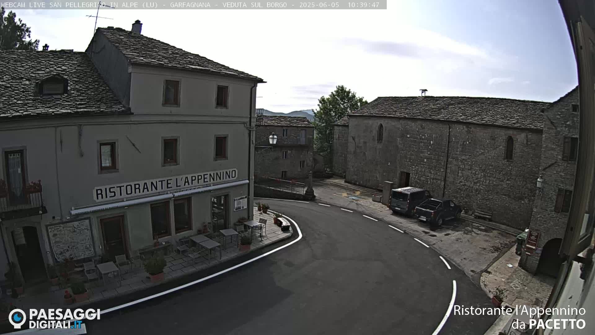 A stone village square with a restaurant and parked vehicles under a mostly clear sky.