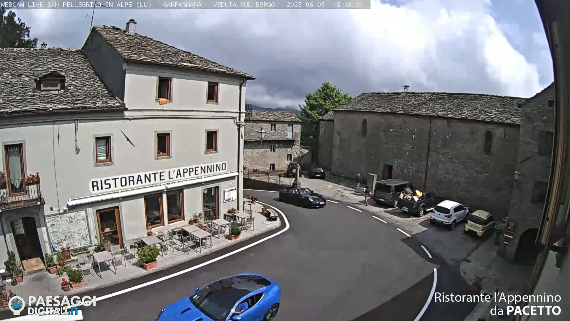 A partly cloudy day reveals a village square with several cars, a restaurant with outdoor seating, and stone buildings.
