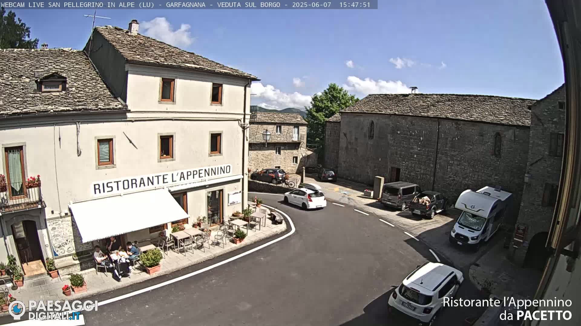 A sunny outdoor scene shows a village square with several cars and a camper van parked near stone buildings, one of which houses a restaurant with outdoor seating.