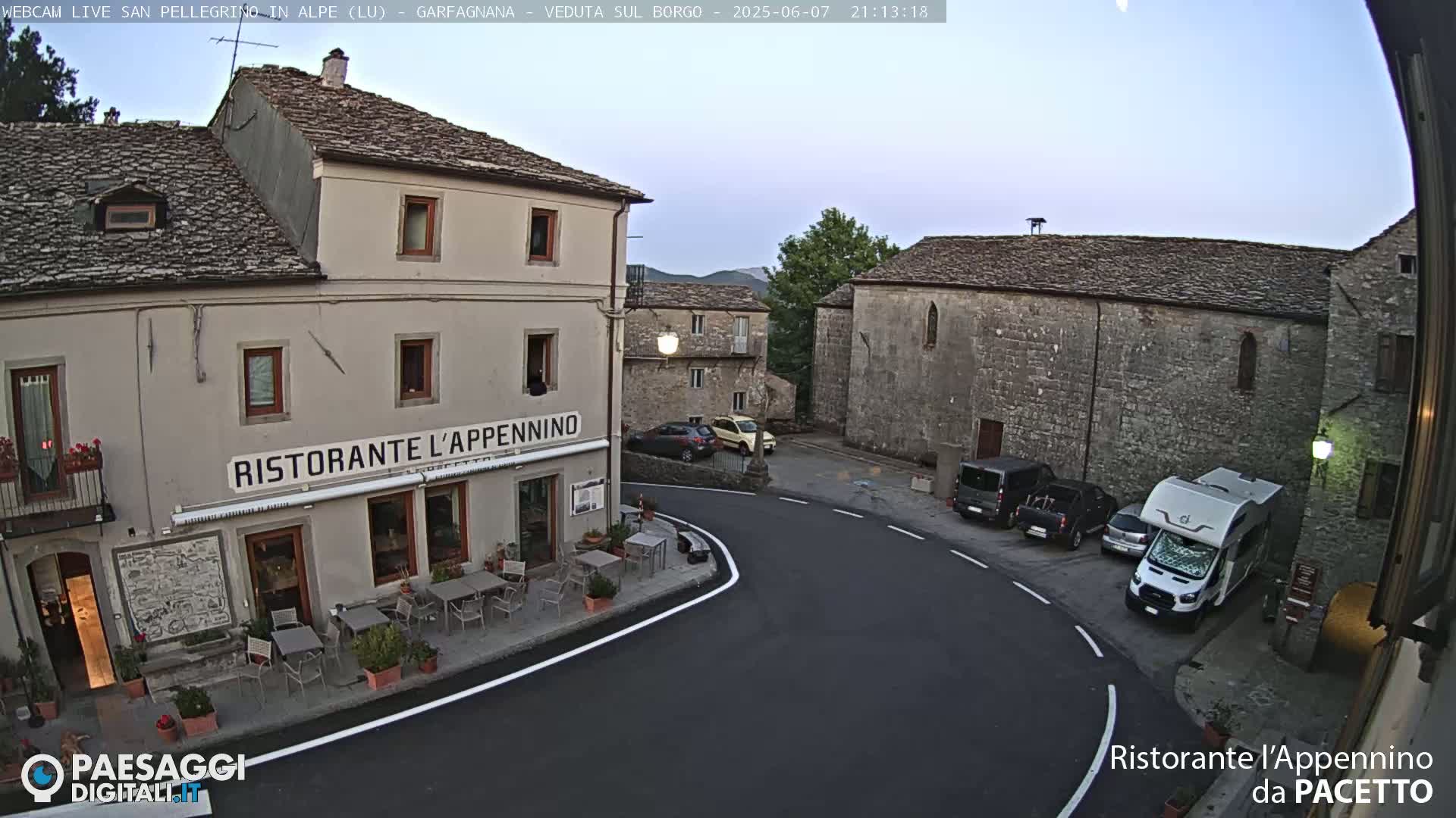 A quaint village square at dusk features a restaurant with outdoor seating, several parked cars and vans, and stone buildings under a clear sky.