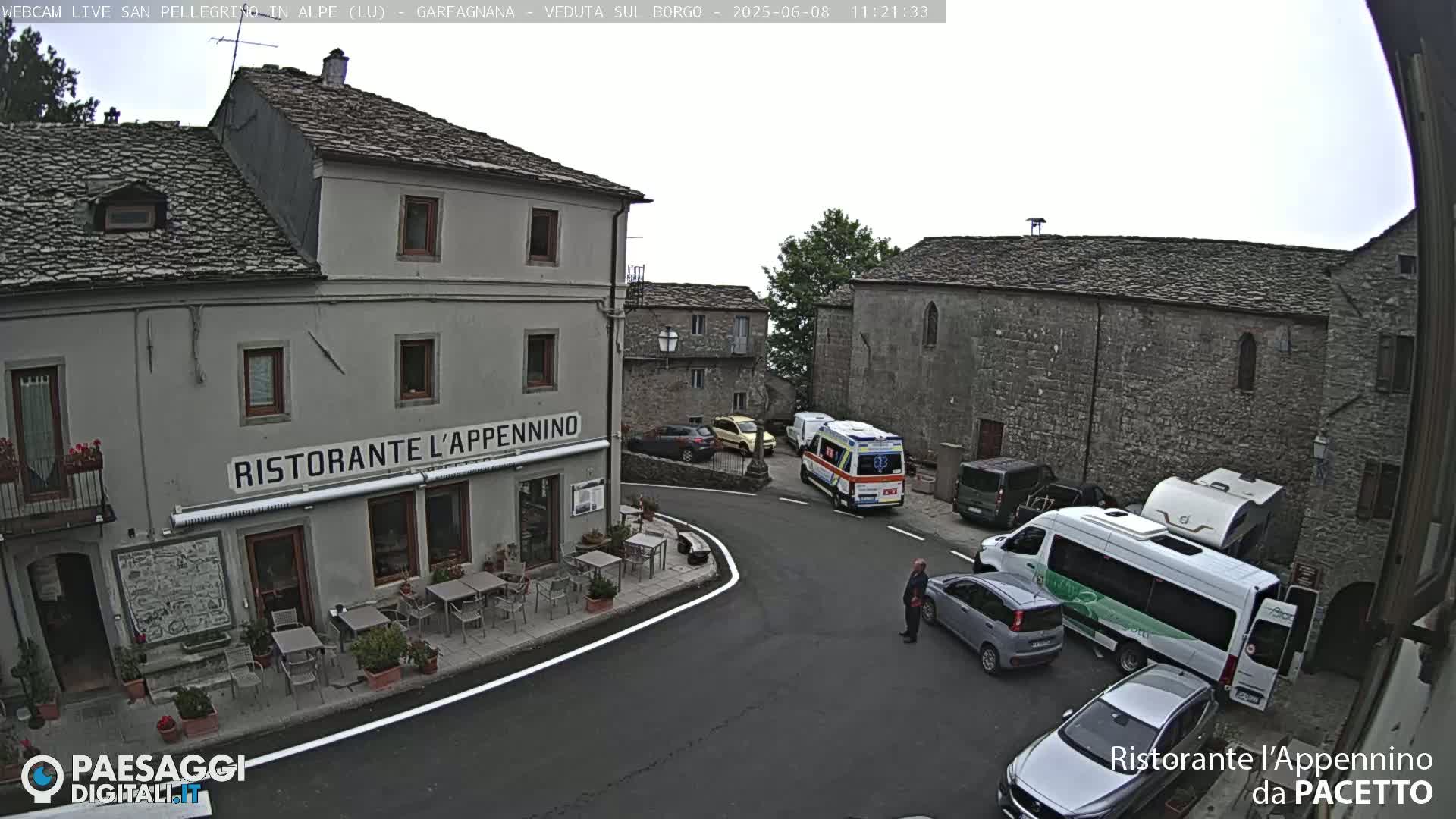 A village square under an overcast sky, with a restaurant, parked cars, a van, and an ambulance visible.