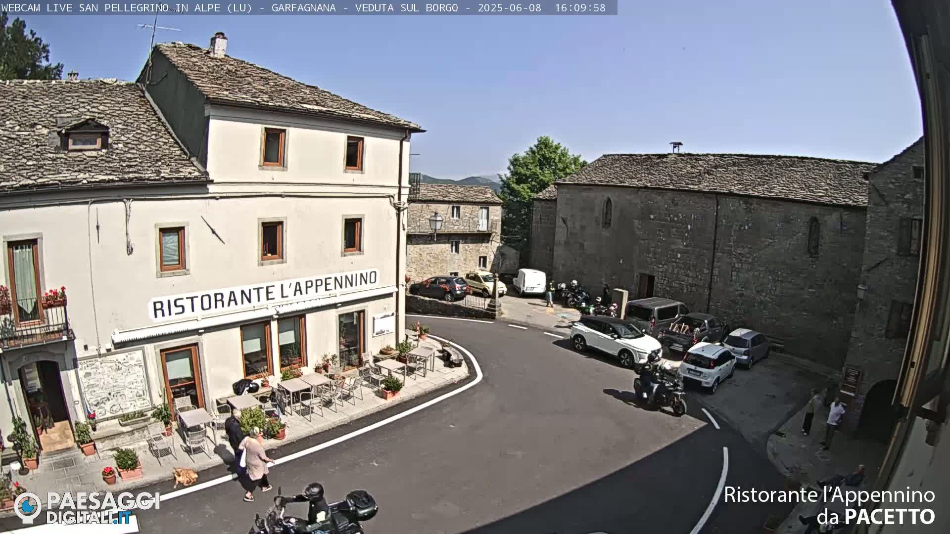 A sunny outdoor scene shows a town square with a restaurant, several parked cars and motorcycles, and a few people walking around.