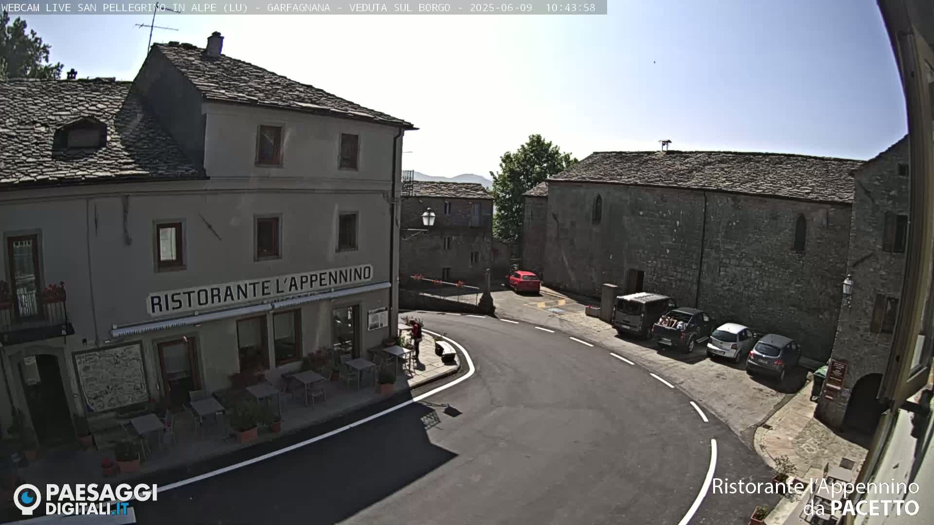 A sunny, outdoor view of a village square with stone buildings, a restaurant, parked cars, and a person standing near a table.