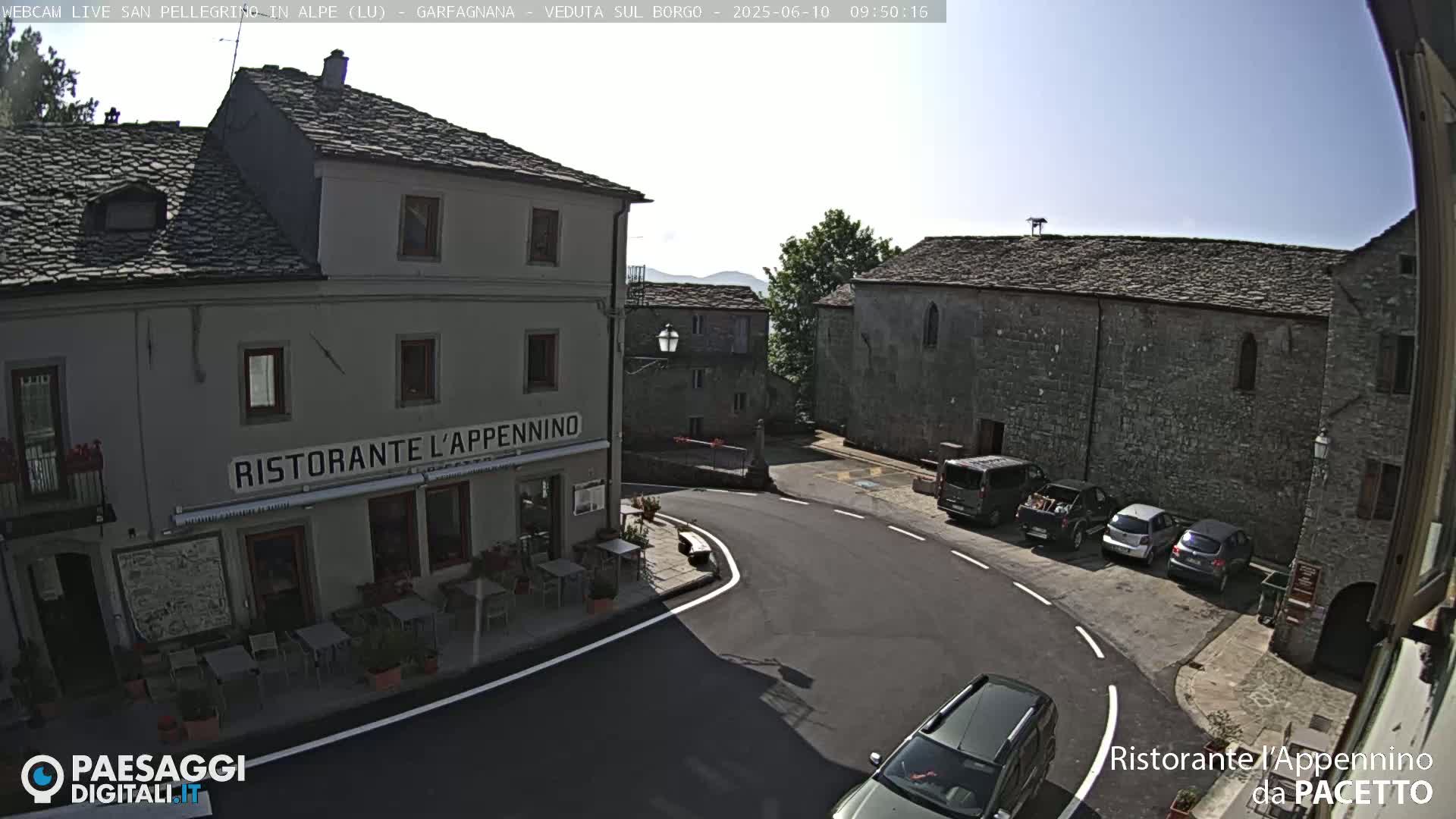 A sunny, outdoor view of a town square with several buildings, cars parked along the side of a street, and a restaurant with outdoor seating.