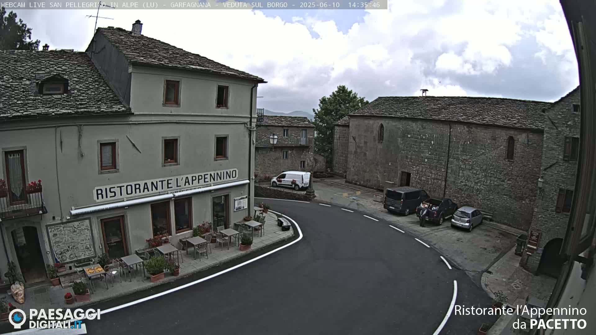 A partly cloudy day reveals a village square with several parked cars, a restaurant with outdoor seating, and stone buildings.