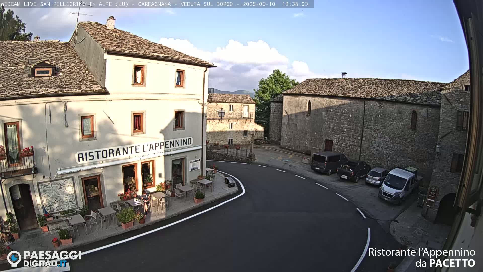 A partly sunny view of a quaint European village square with a restaurant, stone buildings, and parked cars.
