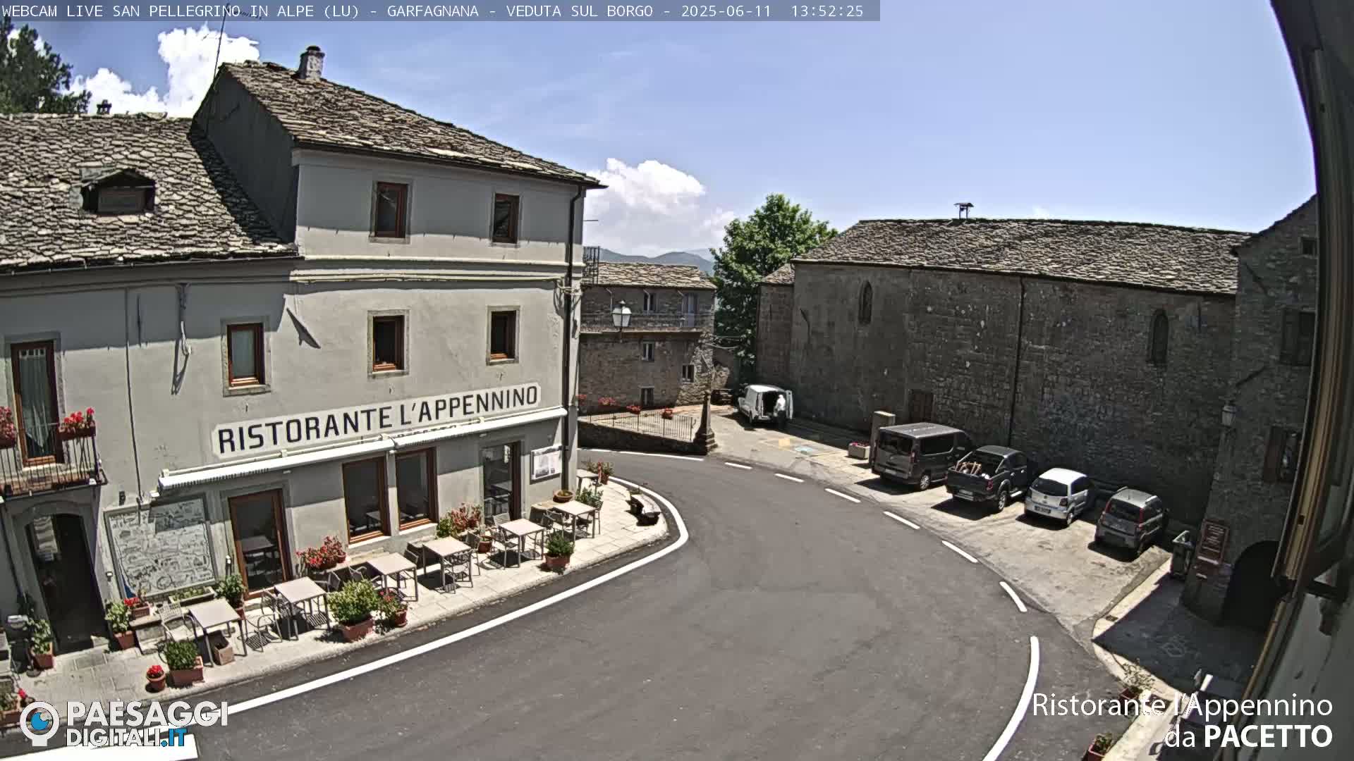 A sunny day shows a village square with several parked cars, a restaurant with outdoor seating, and stone buildings under a mostly clear blue sky.